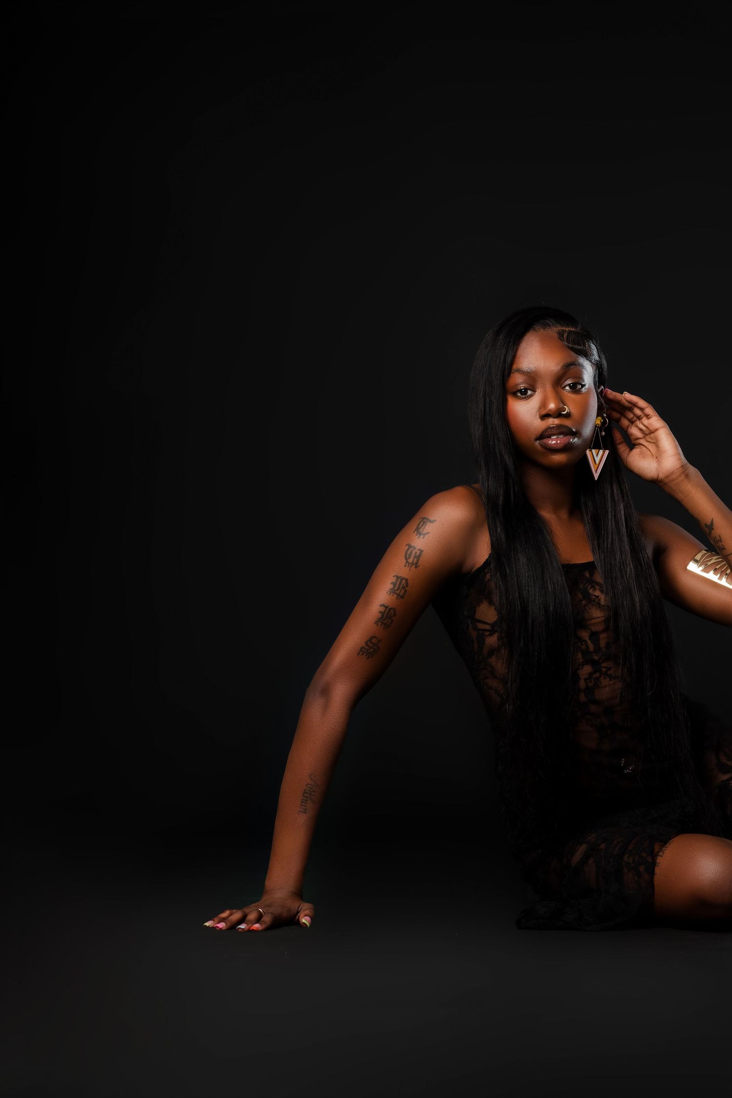 Woman in black lace dress sitting on floor with gold accessories and tattoos on dark background.