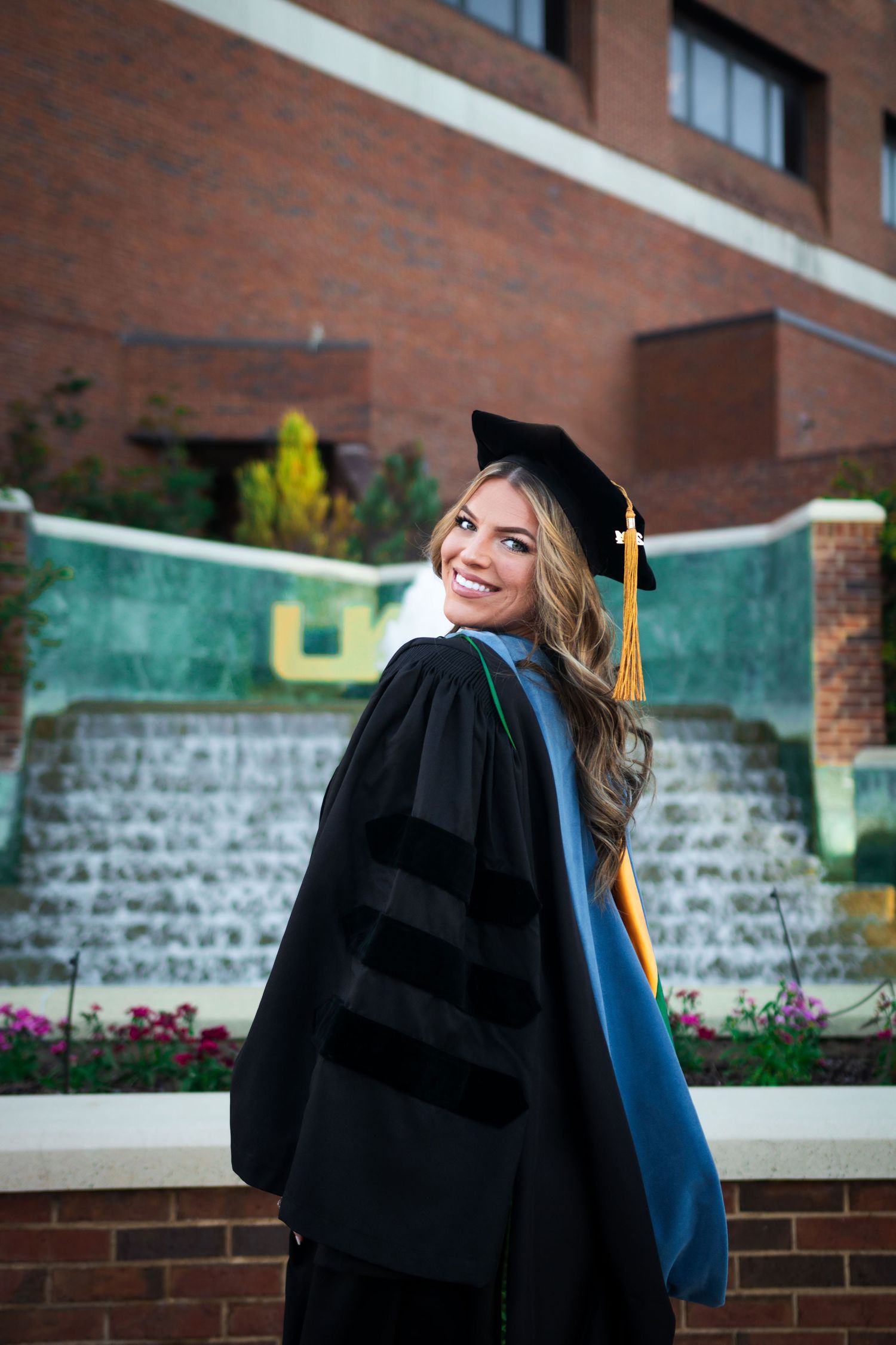 Smiling graduate in doctoral regalia with blue hood turning back toward camera by campus fountain.