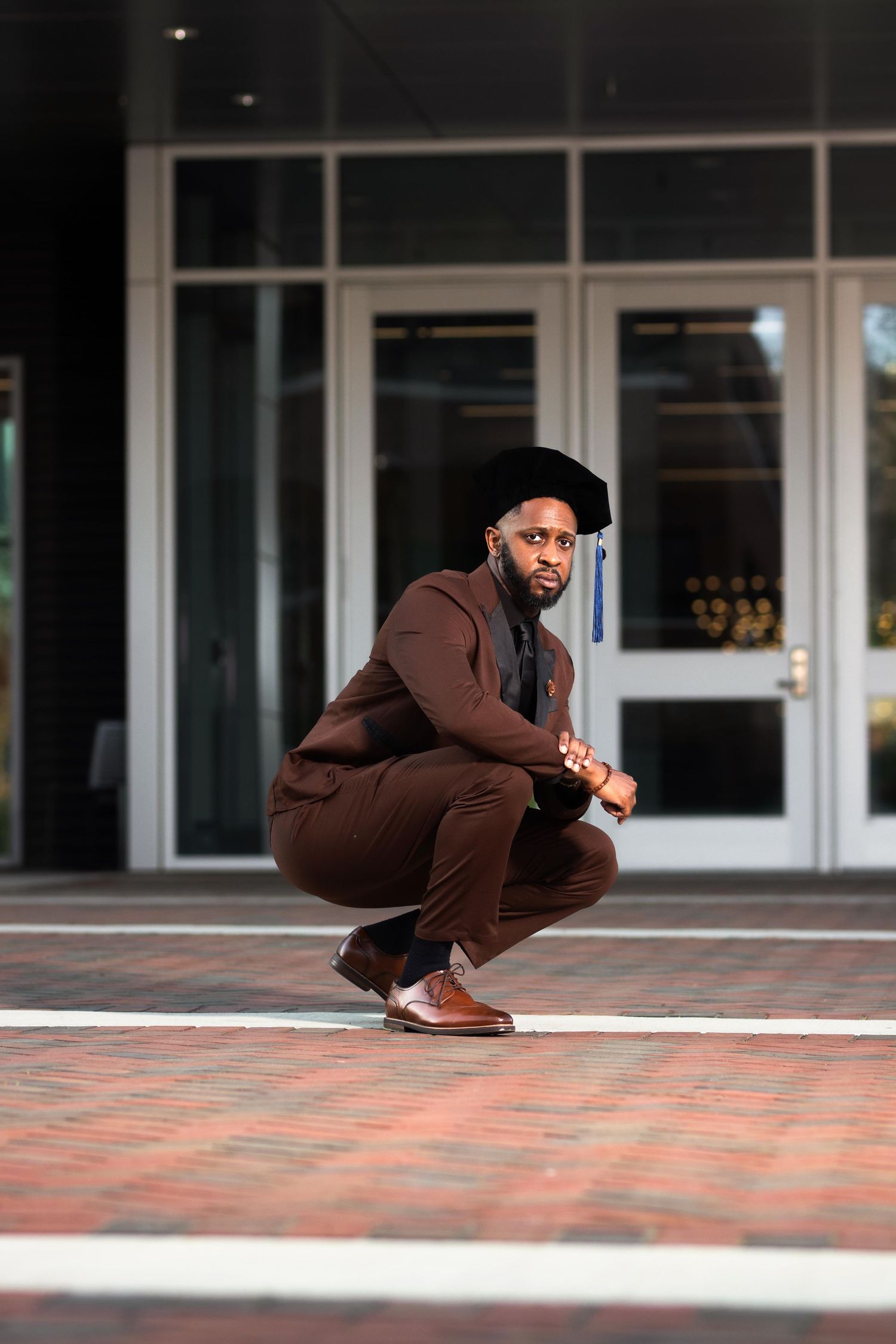 Young Black man in brown suit and doctoral graduation cap crouching outside university building.