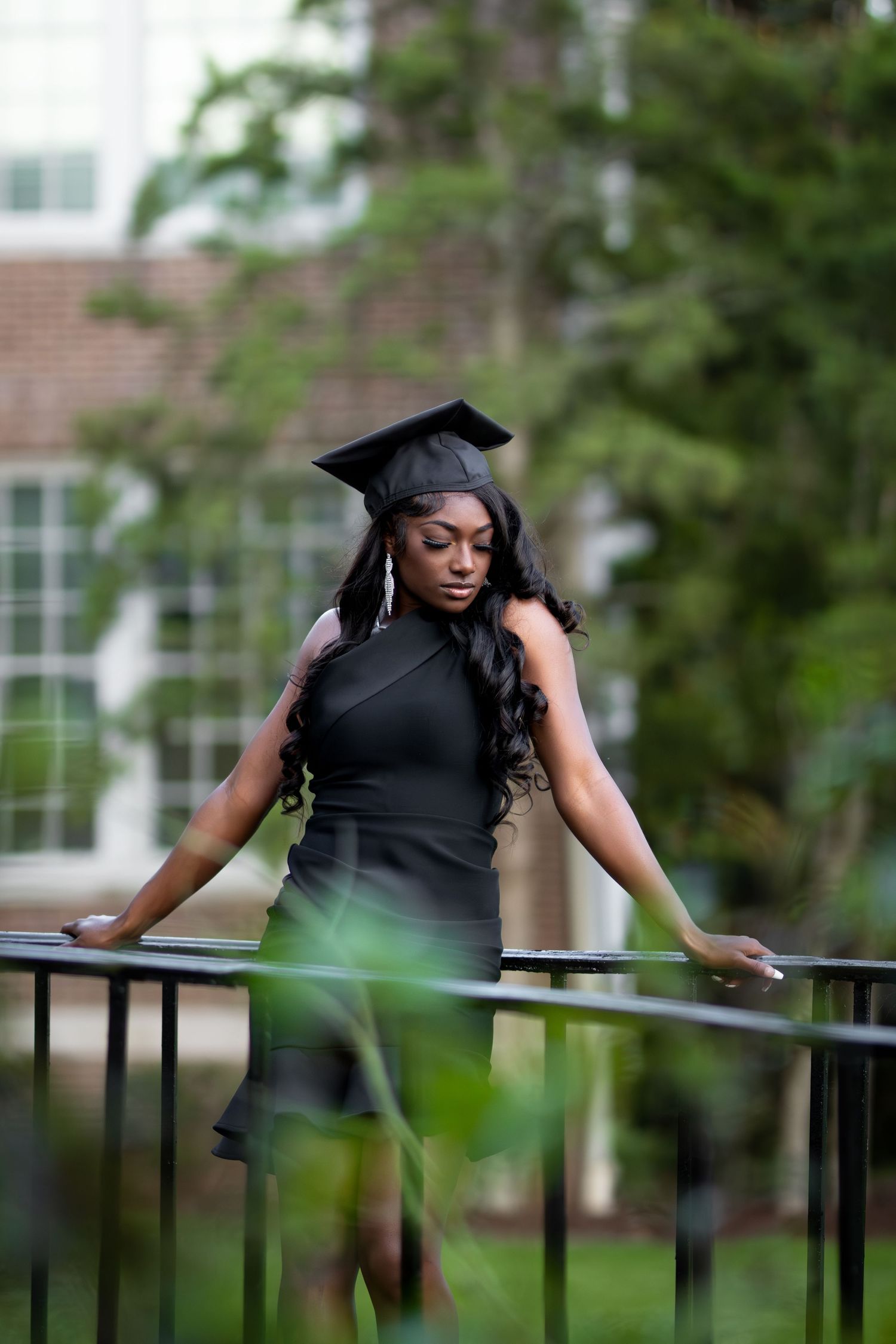 Elegant woman in black dress and graduation cap leans on railing surrounded by lush greenery.