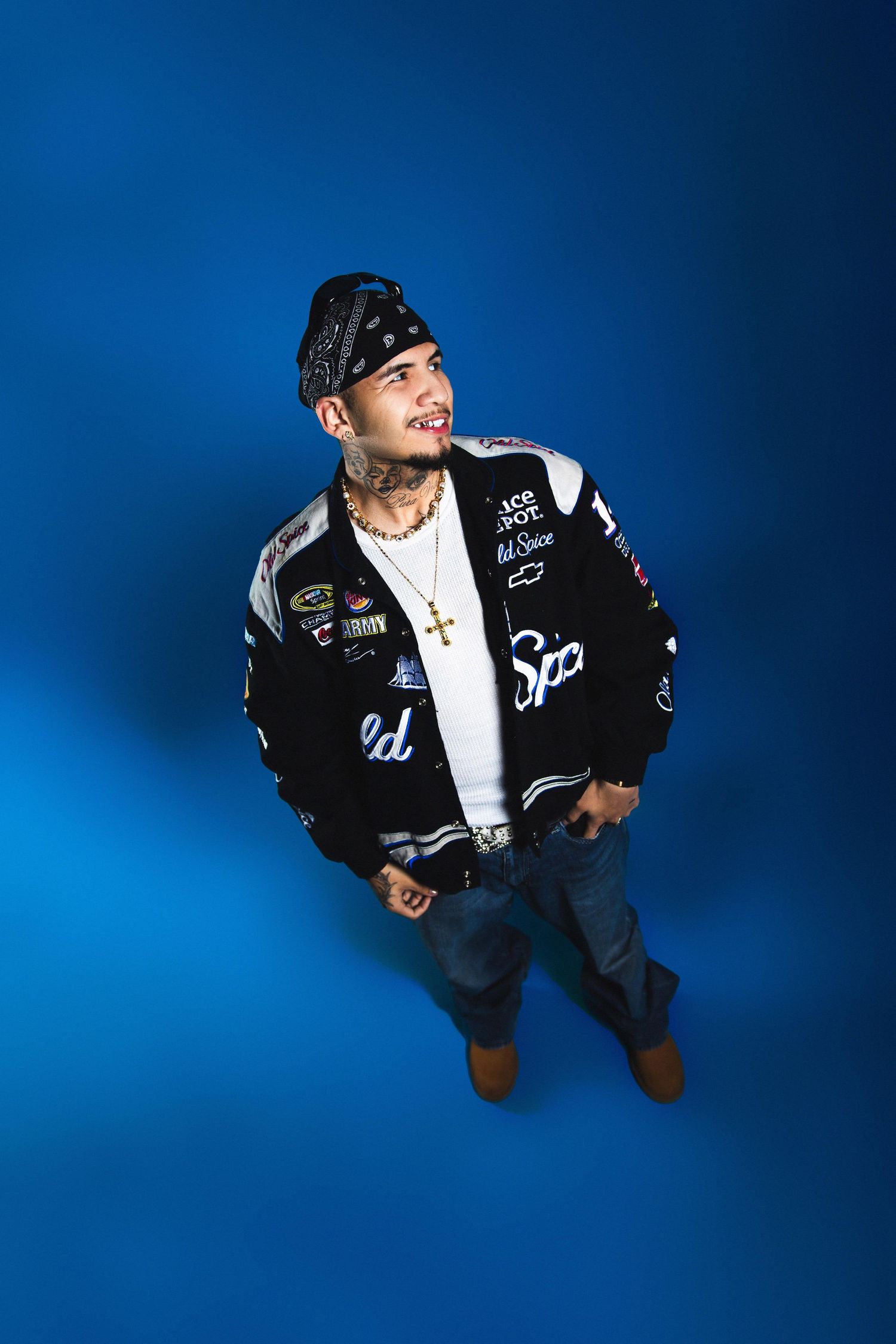 A tattooed man in a bandana and branded racing jacket looks up while posing on a blue background.