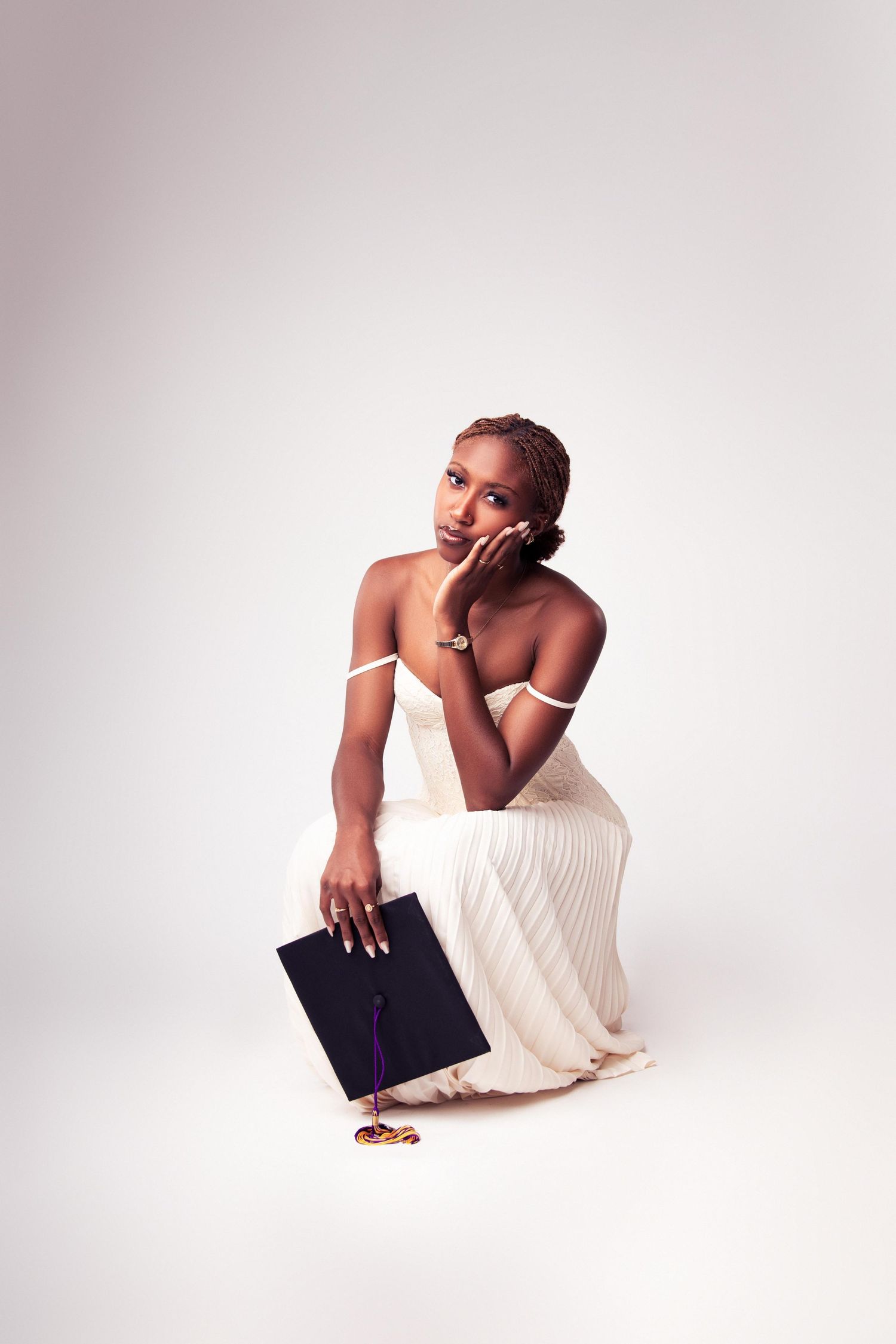 Young woman in white dress holding graduation cap, sitting gracefully in studio portrait photo.