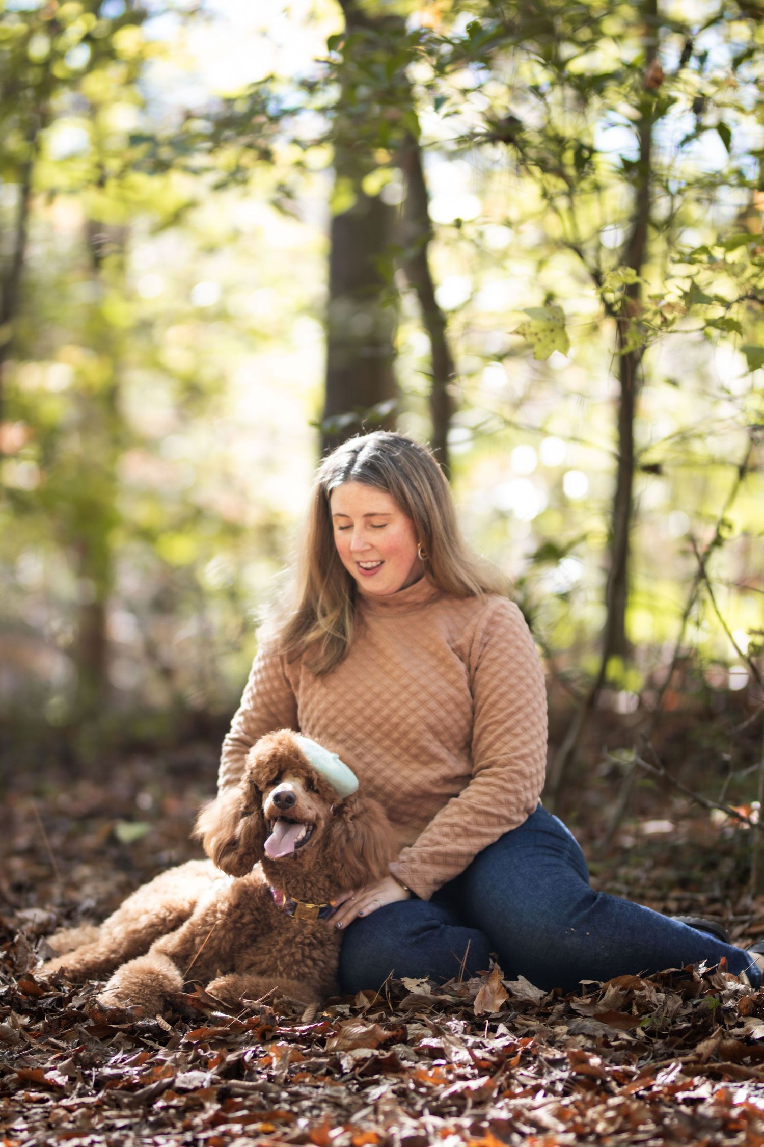 Woman in beige sweater sitting on fallen leaves laughing with her apricot poodle in a sunny forest.