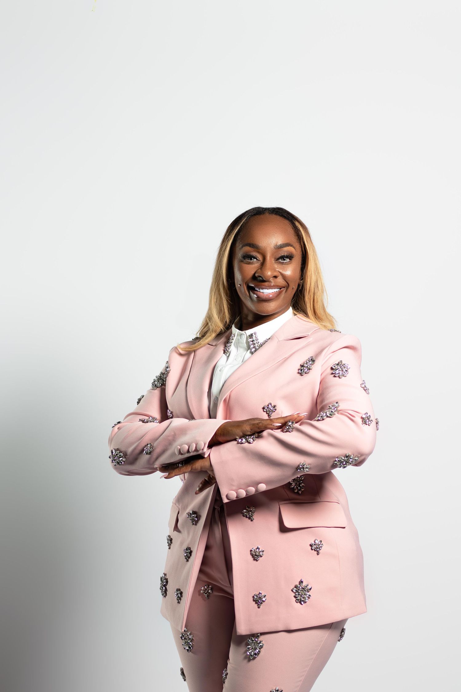 Smiling woman in embellished pink blazer suit with arms crossed posing confidently against white background.
