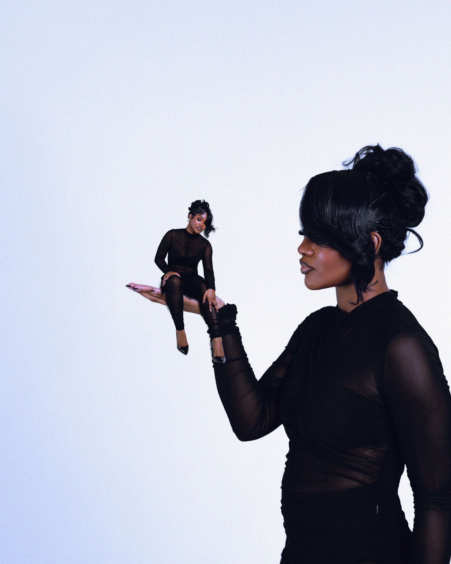 A Black woman in black sheer top holds a tiny doll version of herself against a light background.