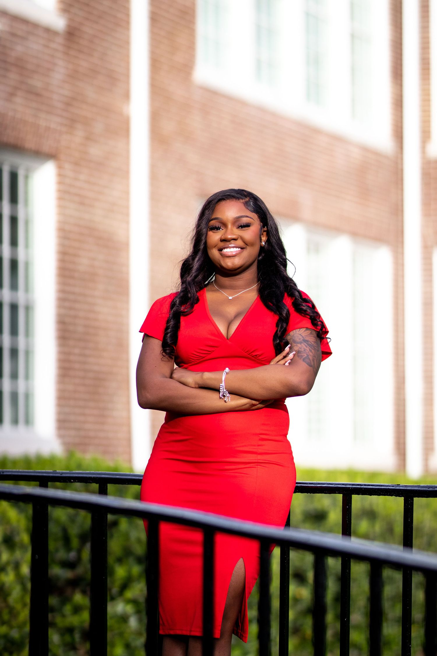 Smiling young woman in red dress stands confidently with arms crossed near brick building.
