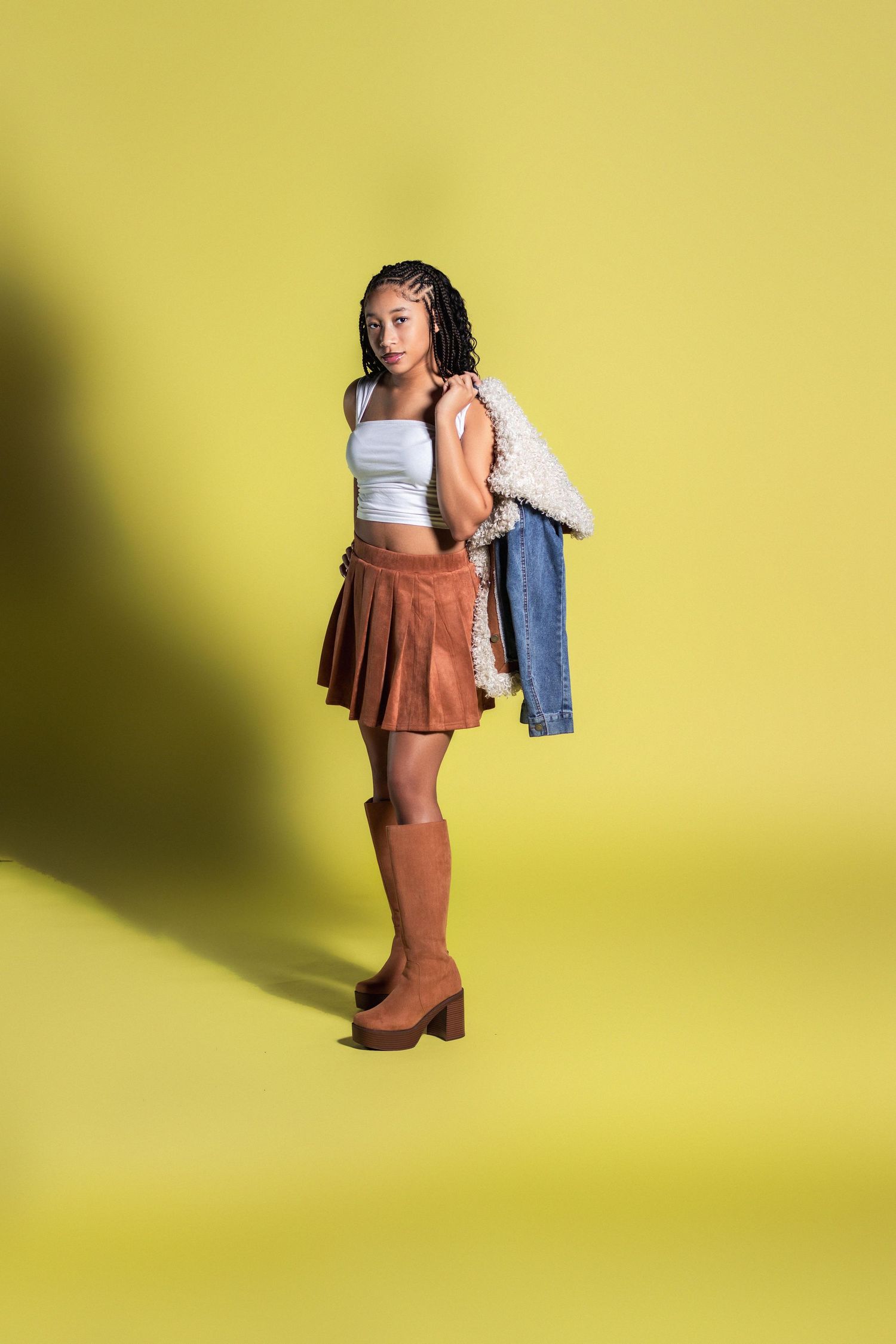 A young Black woman in a brown skirt, white top, and knee-high boots poses on a yellow background.