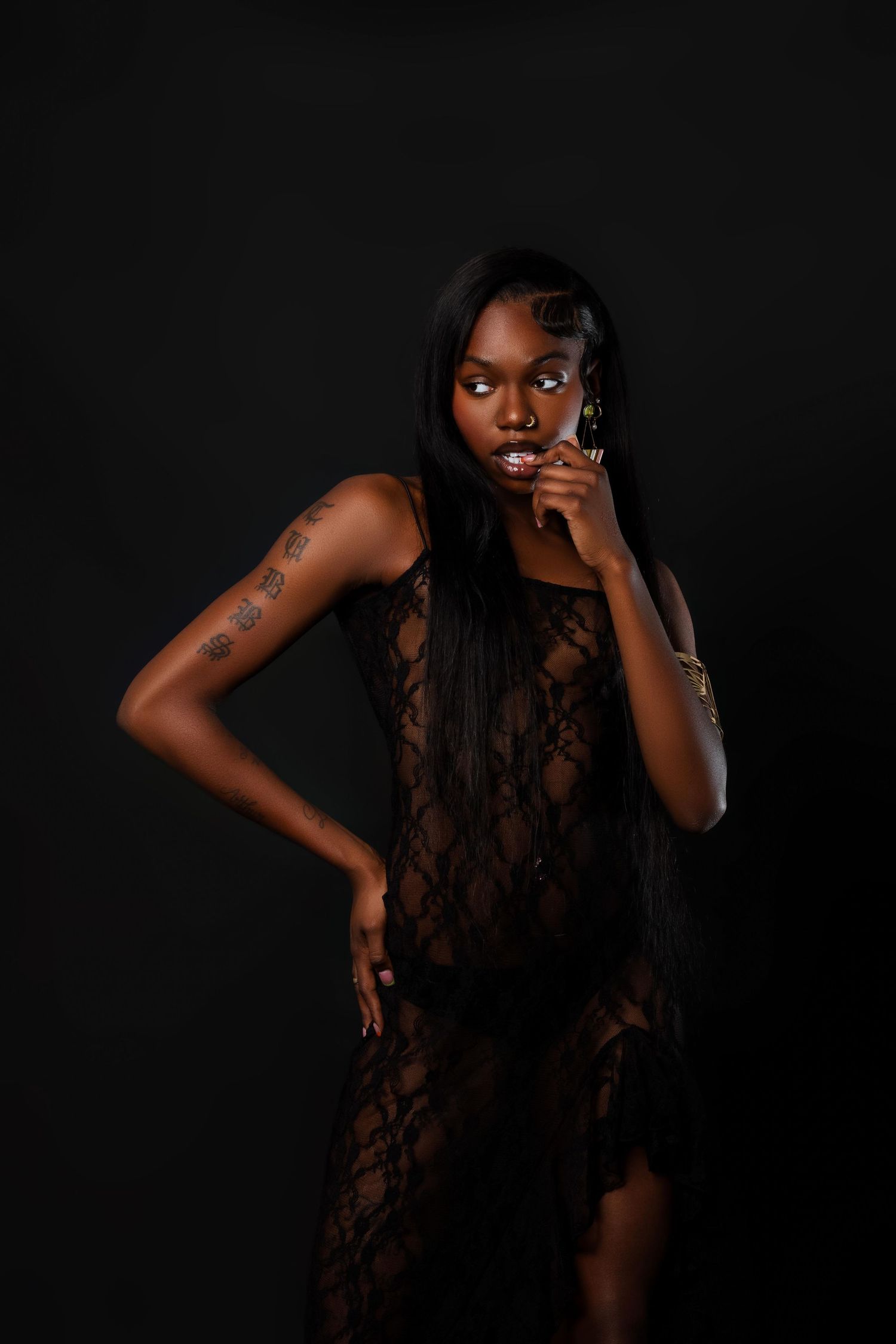 Slender Black woman in black lace dress with tattoos posing against a dark studio background.