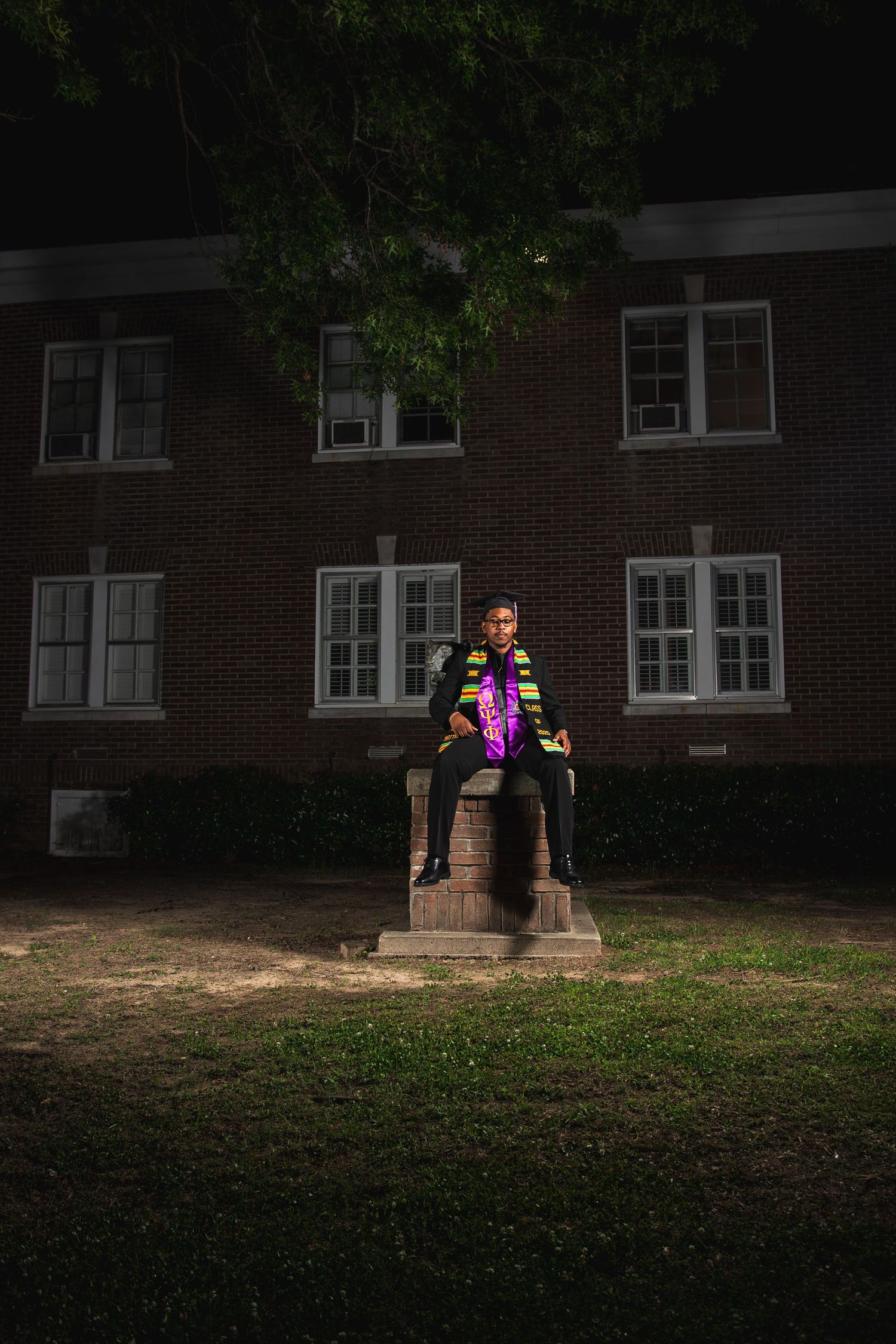 A person in a colorful graduation stole sits atop a stone pedestal at night in front of a brick building.