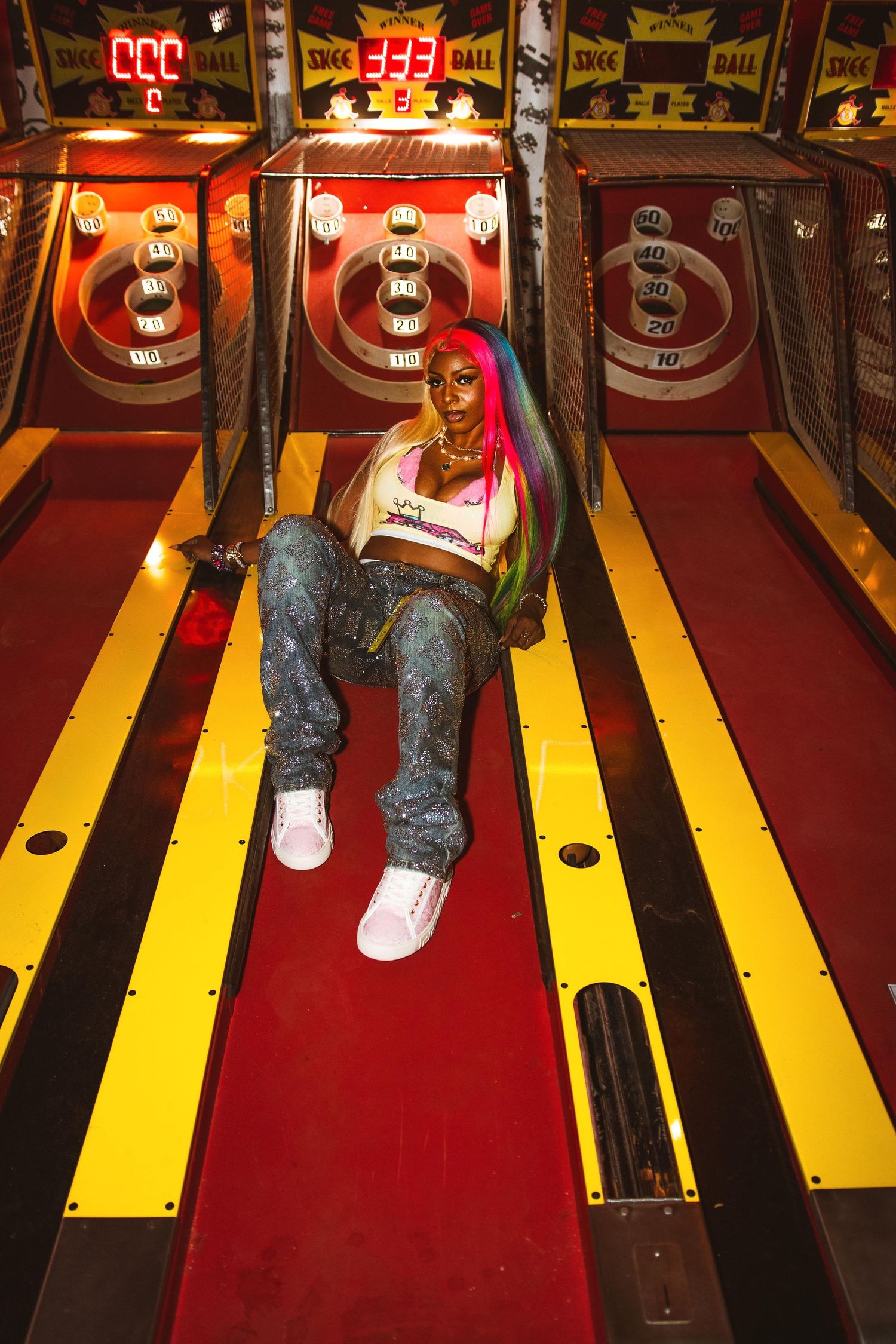 Woman with rainbow hair sits on skee-ball arcade lane in jeans and white top.