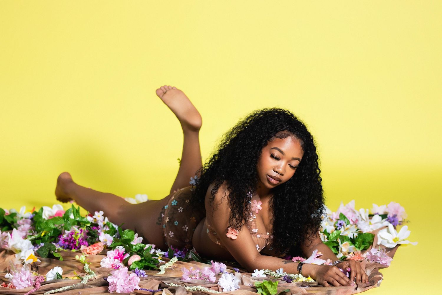 Young woman with curly hair lying among colorful flowers on fabric against yellow background.