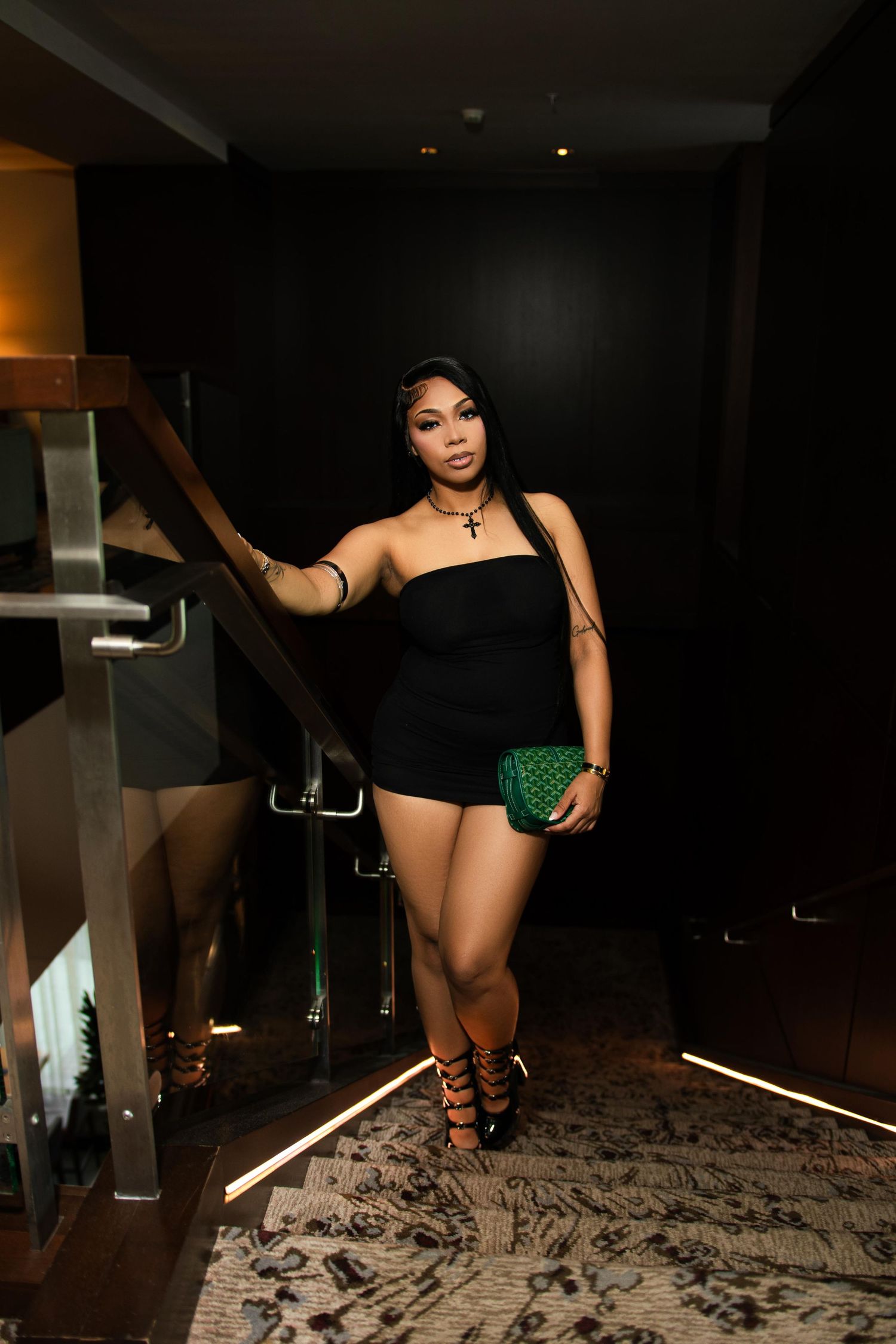 A woman in a black strapless romper holds a green clutch while posing on stairs in a dimly lit venue.