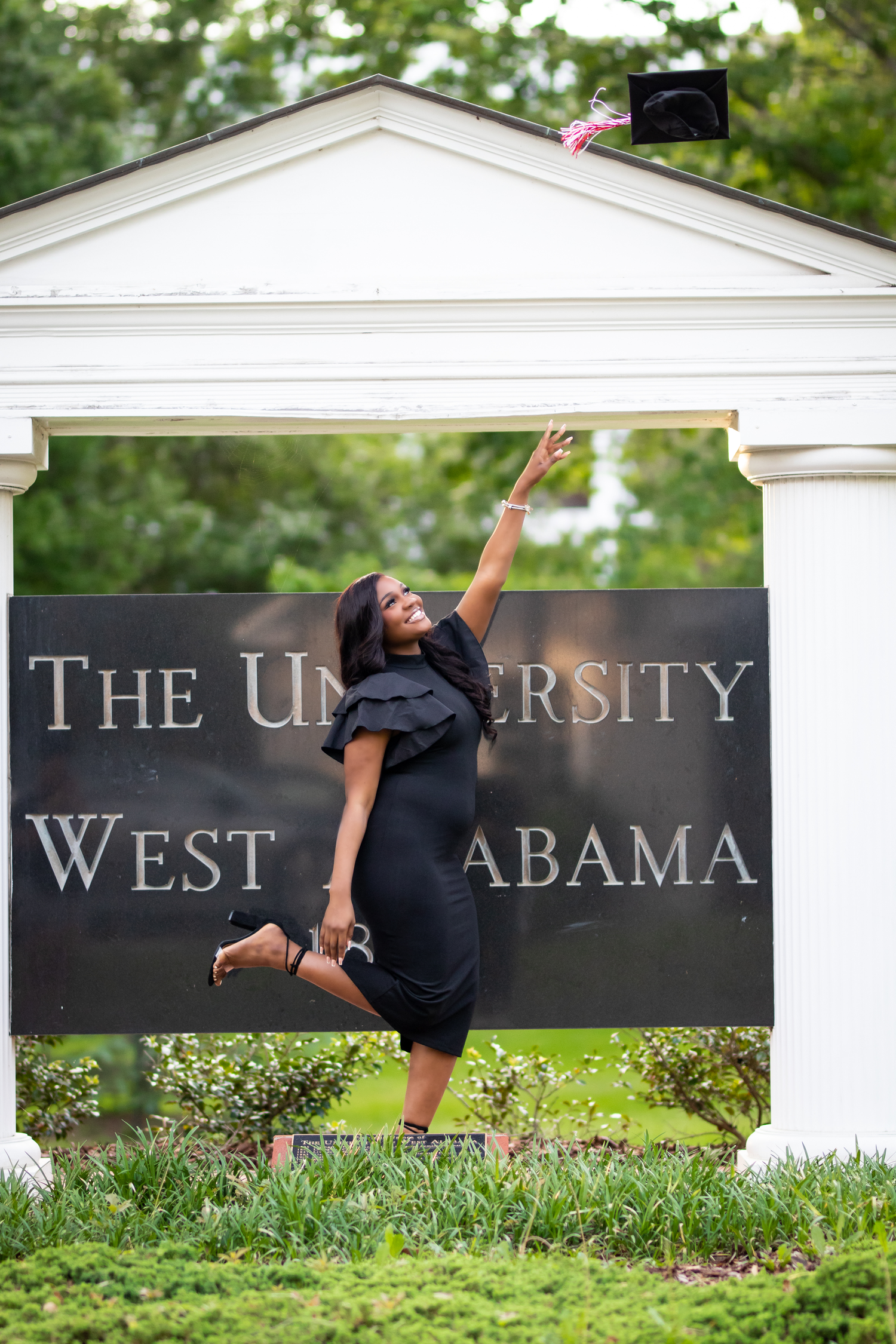 Joyful graduate in black dress tosses cap at University of West Alabama entrance sign.