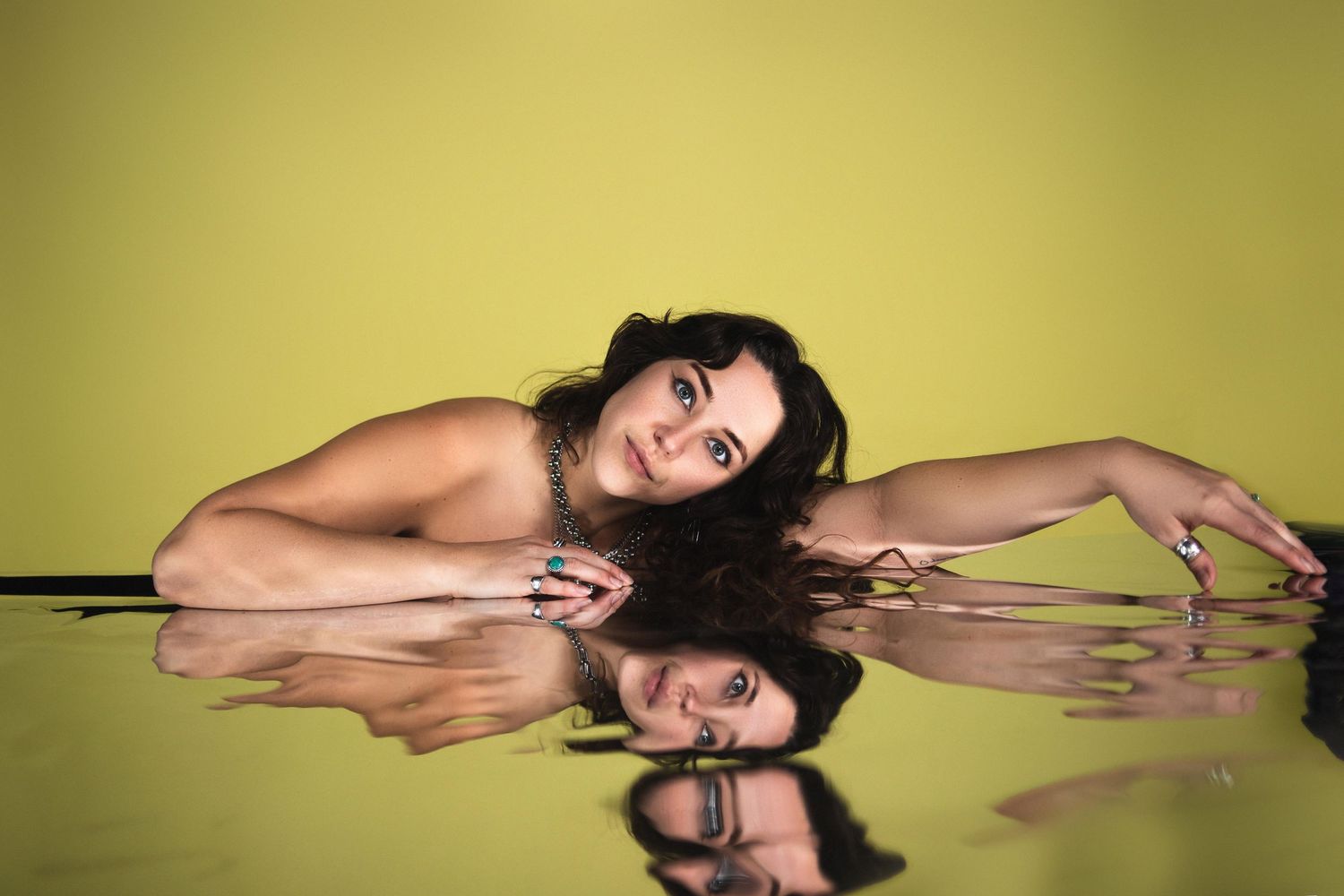 Woman with dark hair lying on reflective surface against yellow-green background with mirror reflection below.