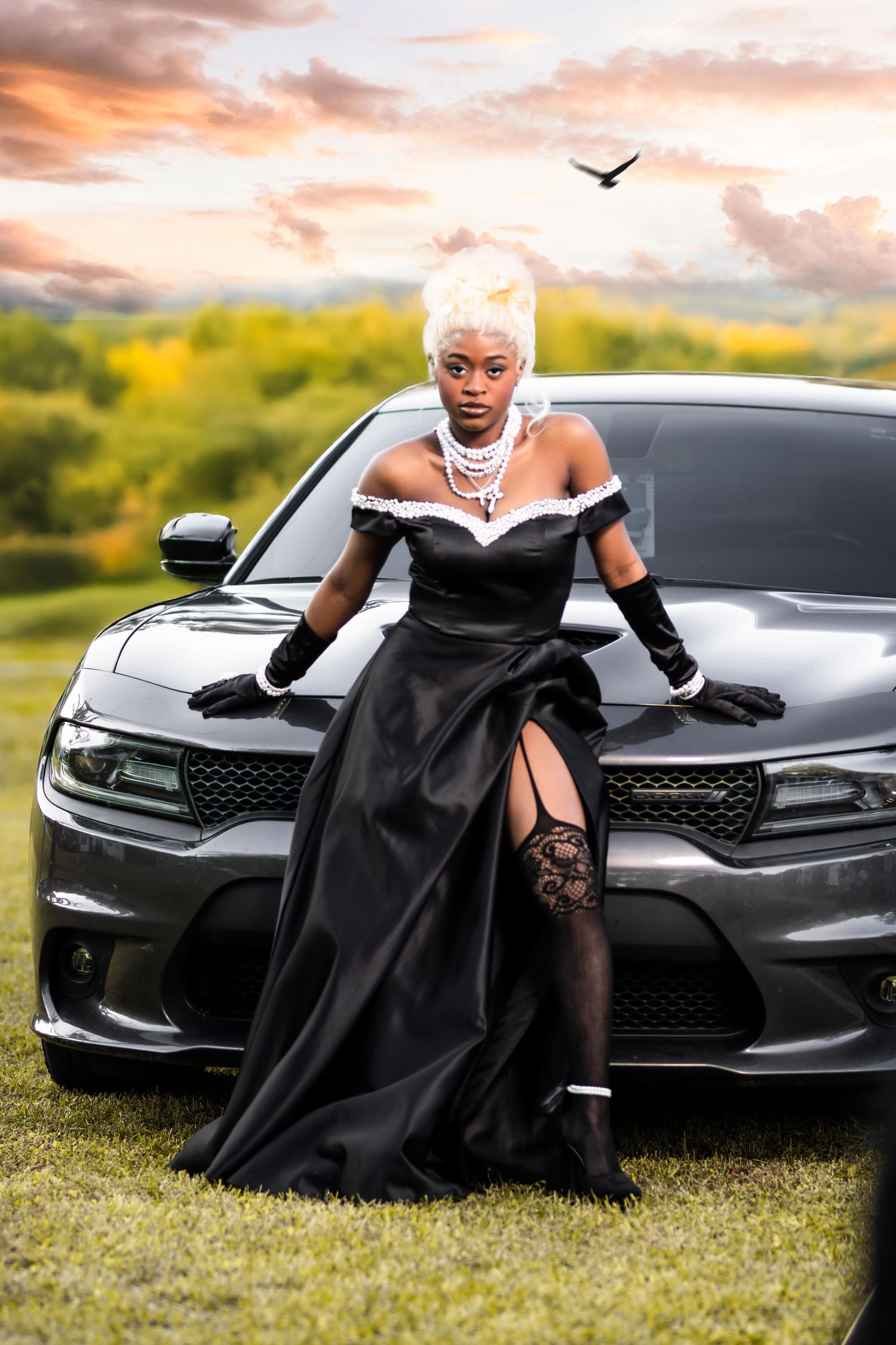 Woman in elegant black off-shoulder gown with white hair poses against black car at sunset.
