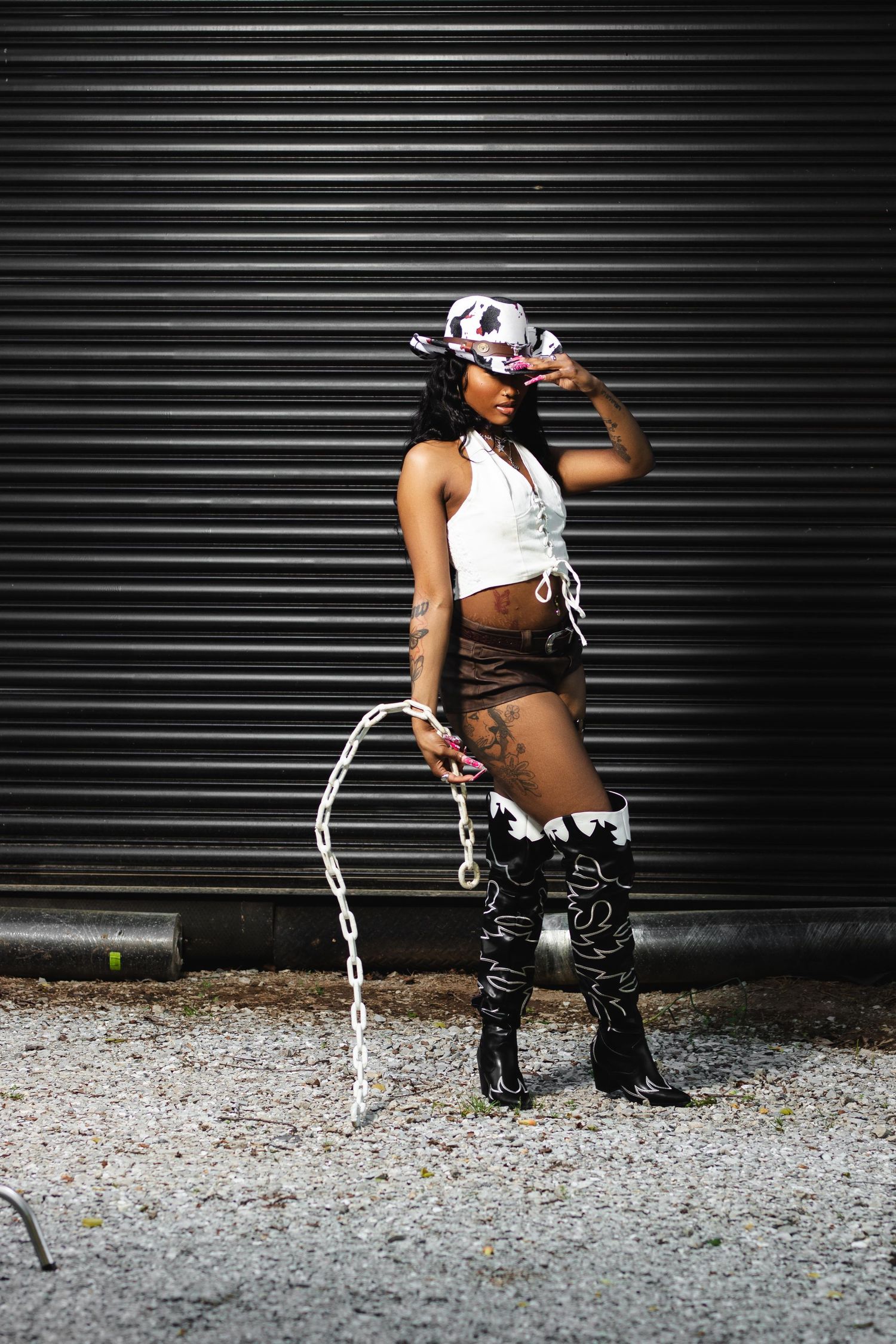 Black woman in cowgirl outfit with hat, fringe skirt, and boots holding a whip against a metal shutter backdrop.