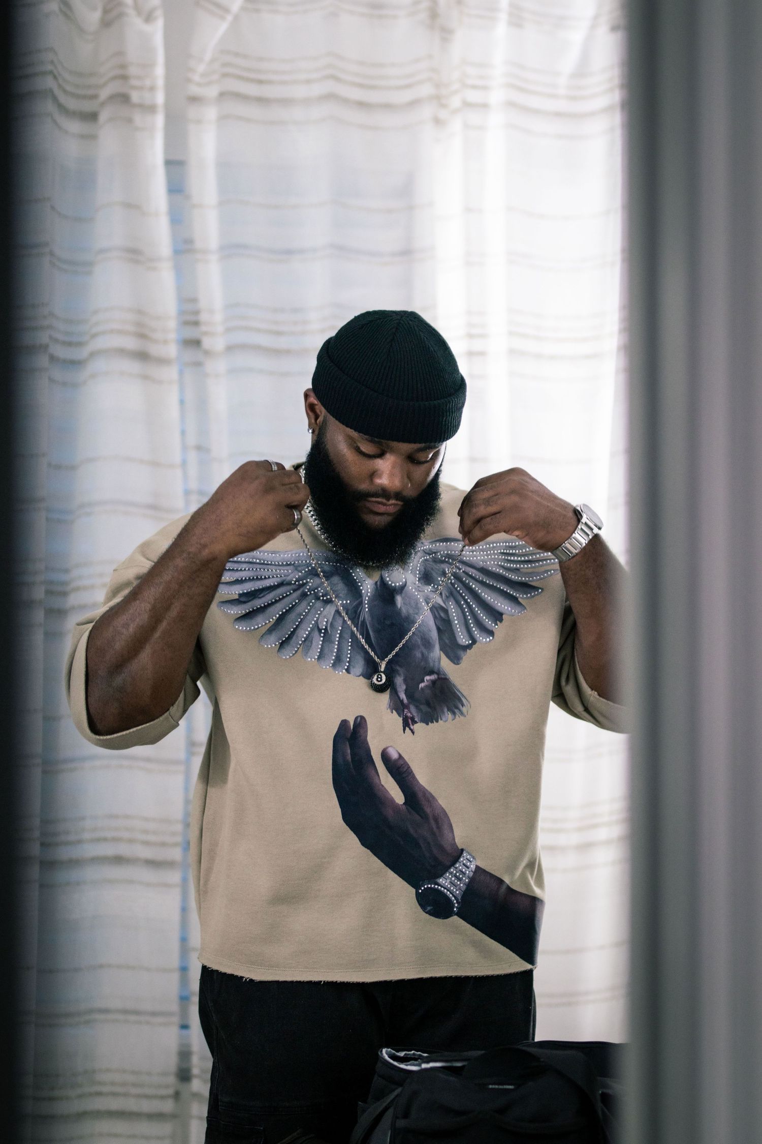 Man in beige graphic bird t-shirt and black beanie adjusting collar indoors.