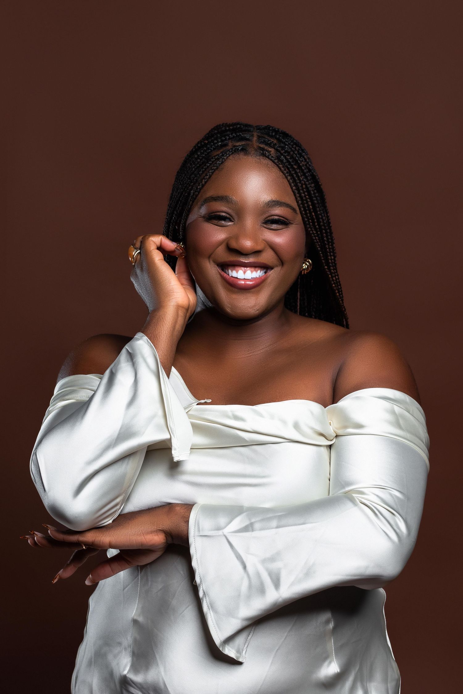 Smiling Black woman in white off-shoulder satin dress with braids, posing against brown background.