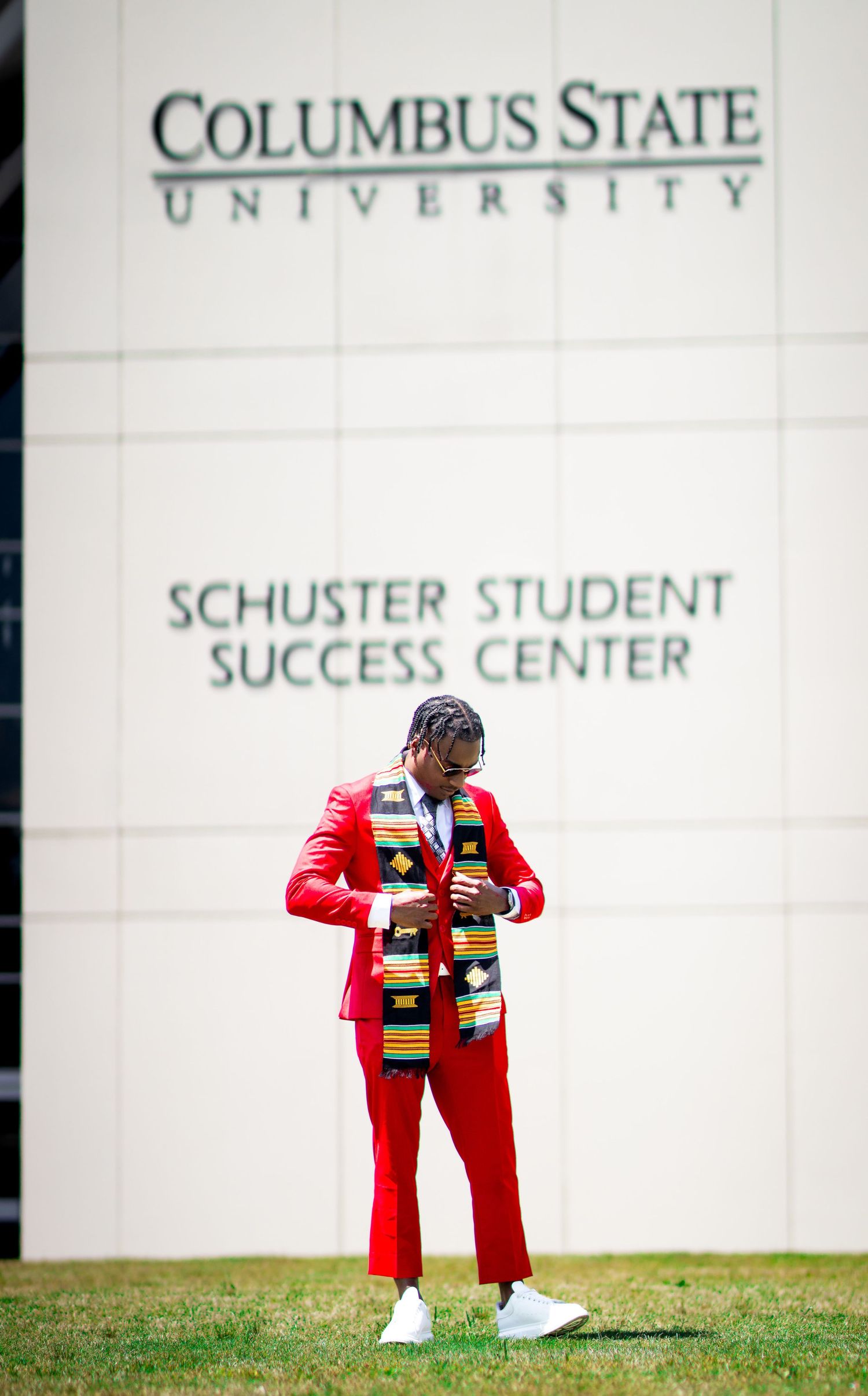 Graduate in red suit with kente stole stands before Columbus State University Schuster Student Success Center.
