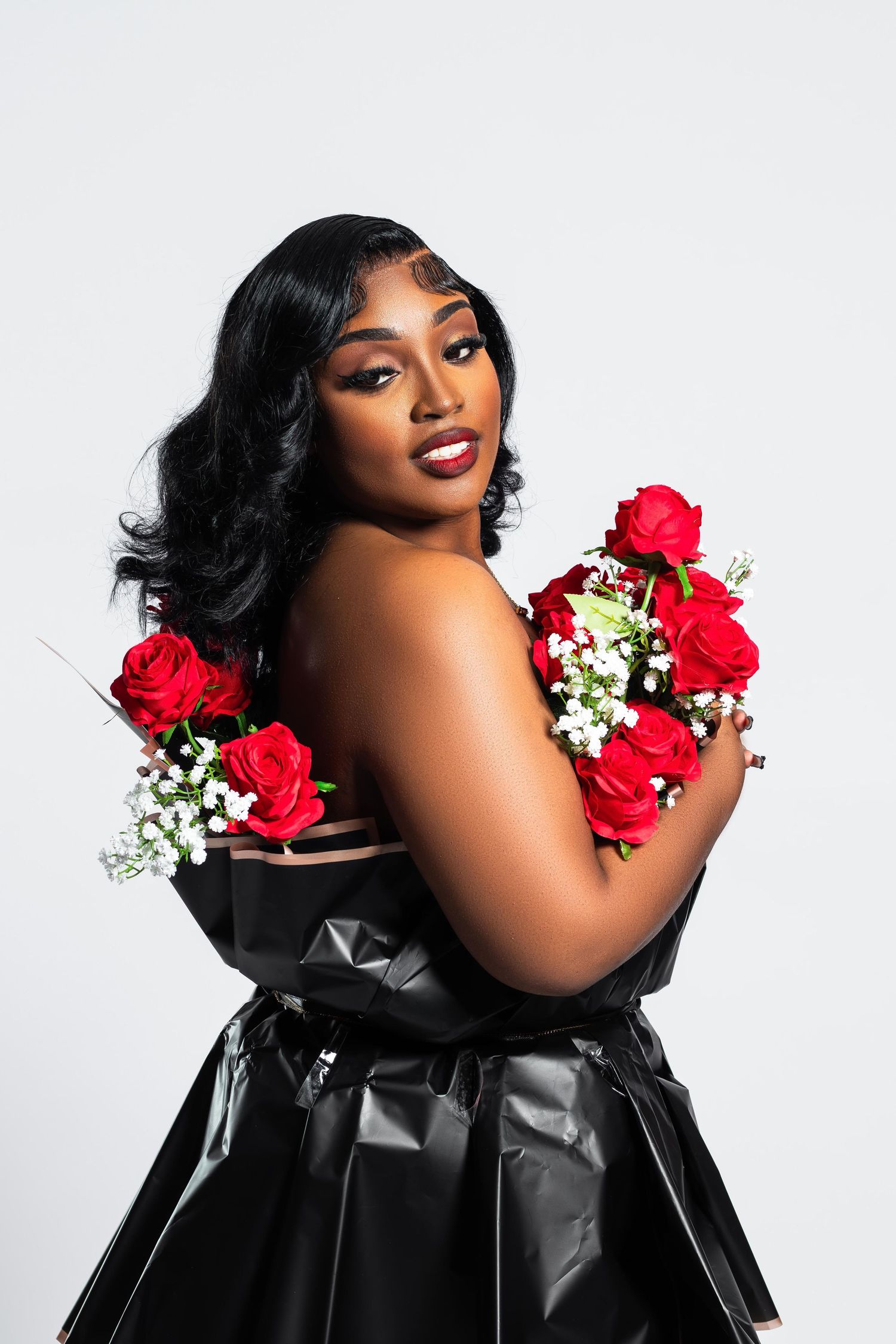 A Black woman in black latex dress poses with red roses and white flowers on gray background.