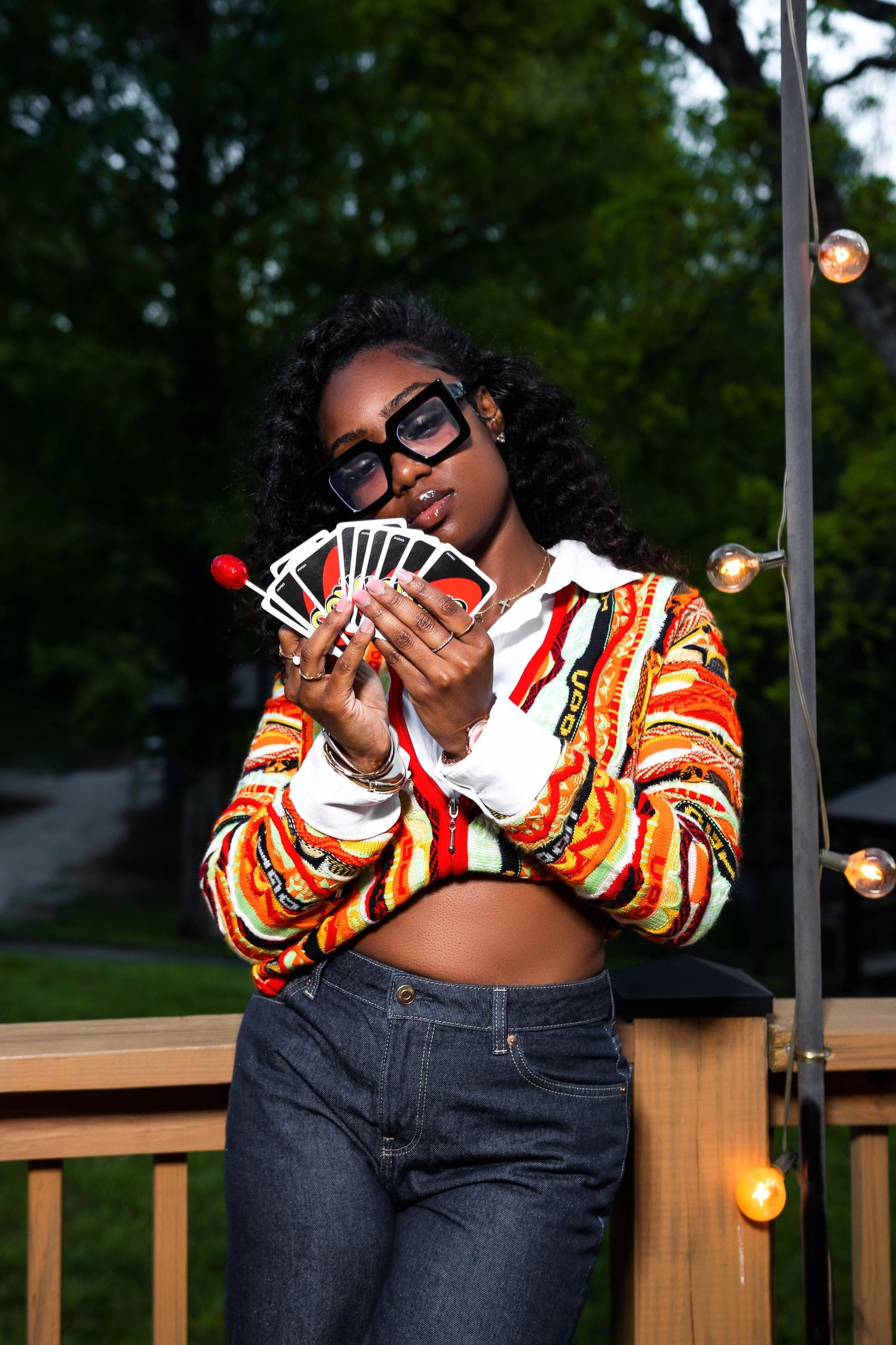 Young Black woman in colorful striped crop jacket and sunglasses holding playing cards on an outdoor deck.