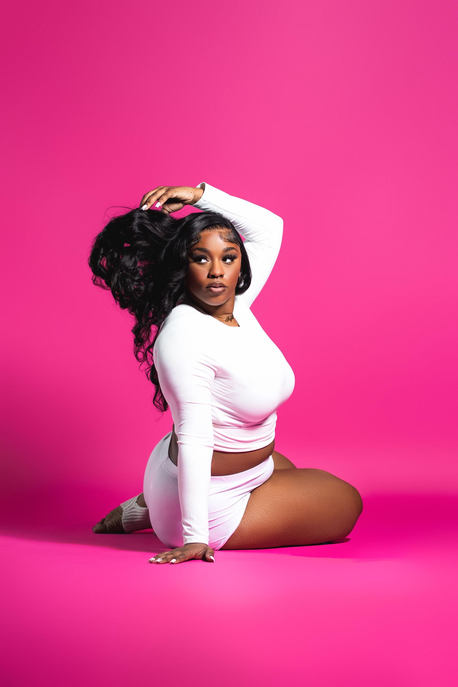A curvy Black woman in a white crop top sits gracefully on a pink studio backdrop with long wavy hair.