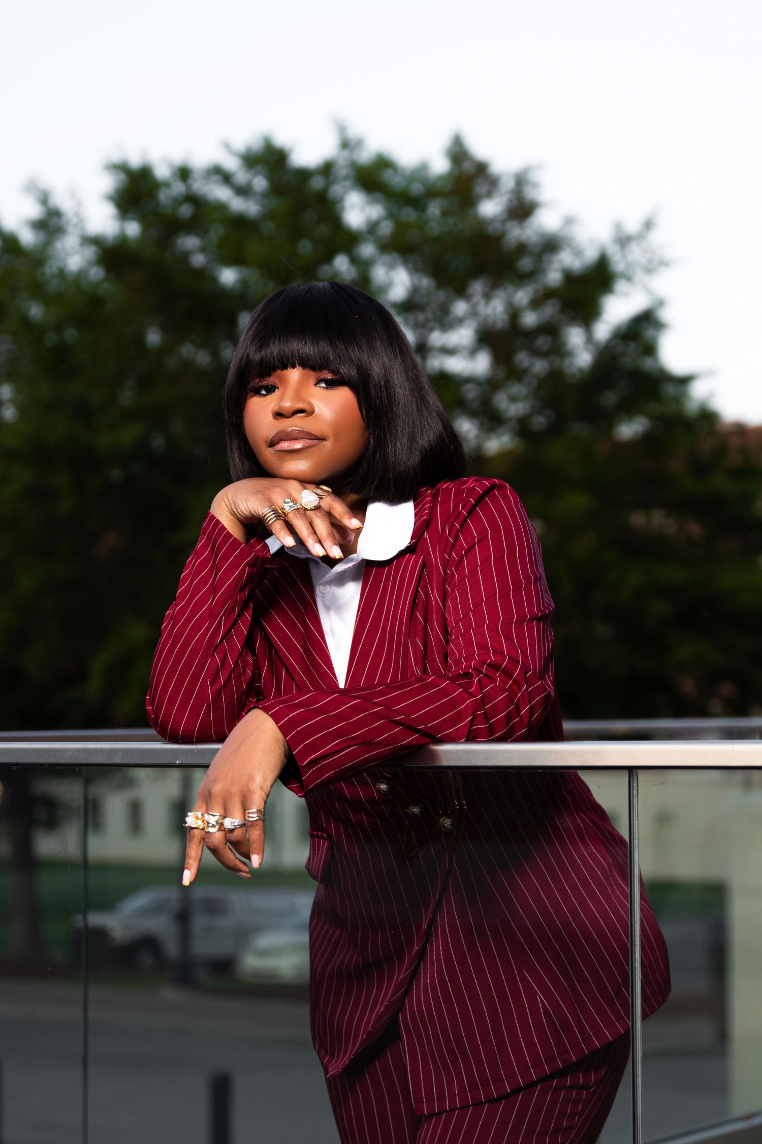 A confident Black woman in a red pinstripe suit leans on a railing outdoors, wearing statement rings.