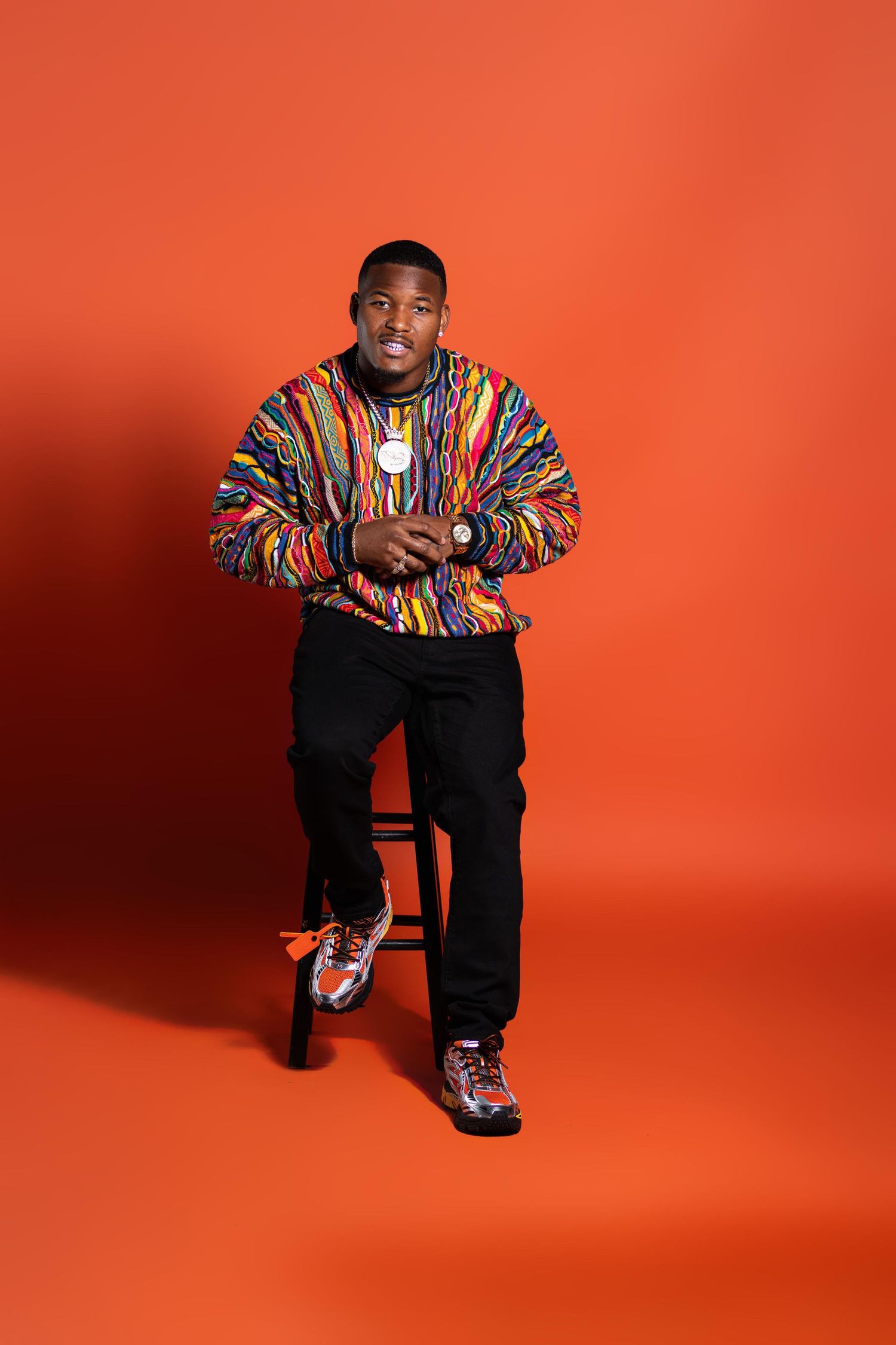 Man in colorful striped jacket sitting on stool against orange background.