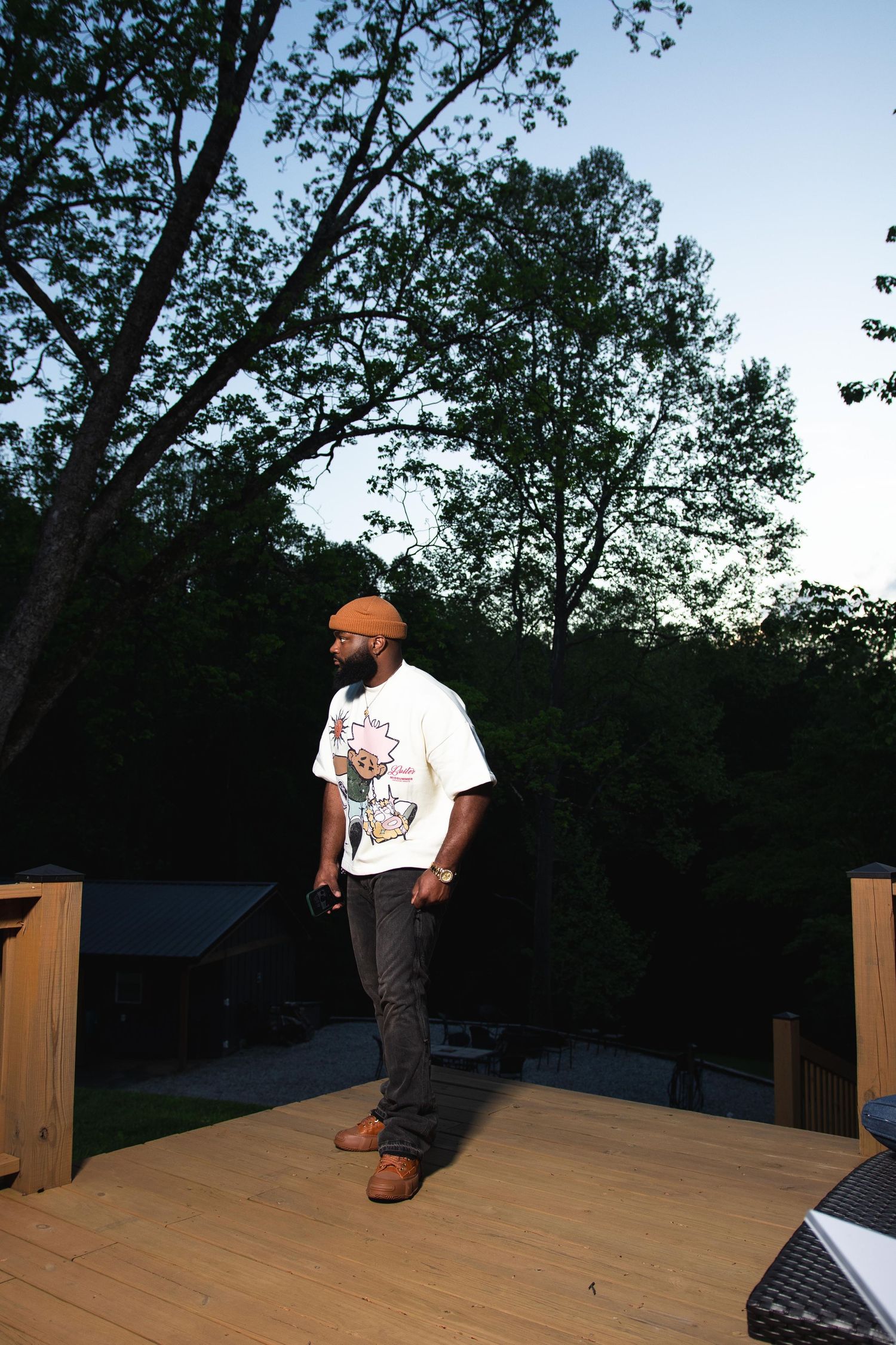 Man in white graphic tee and orange beanie stands on wooden deck at dusk with trees behind.