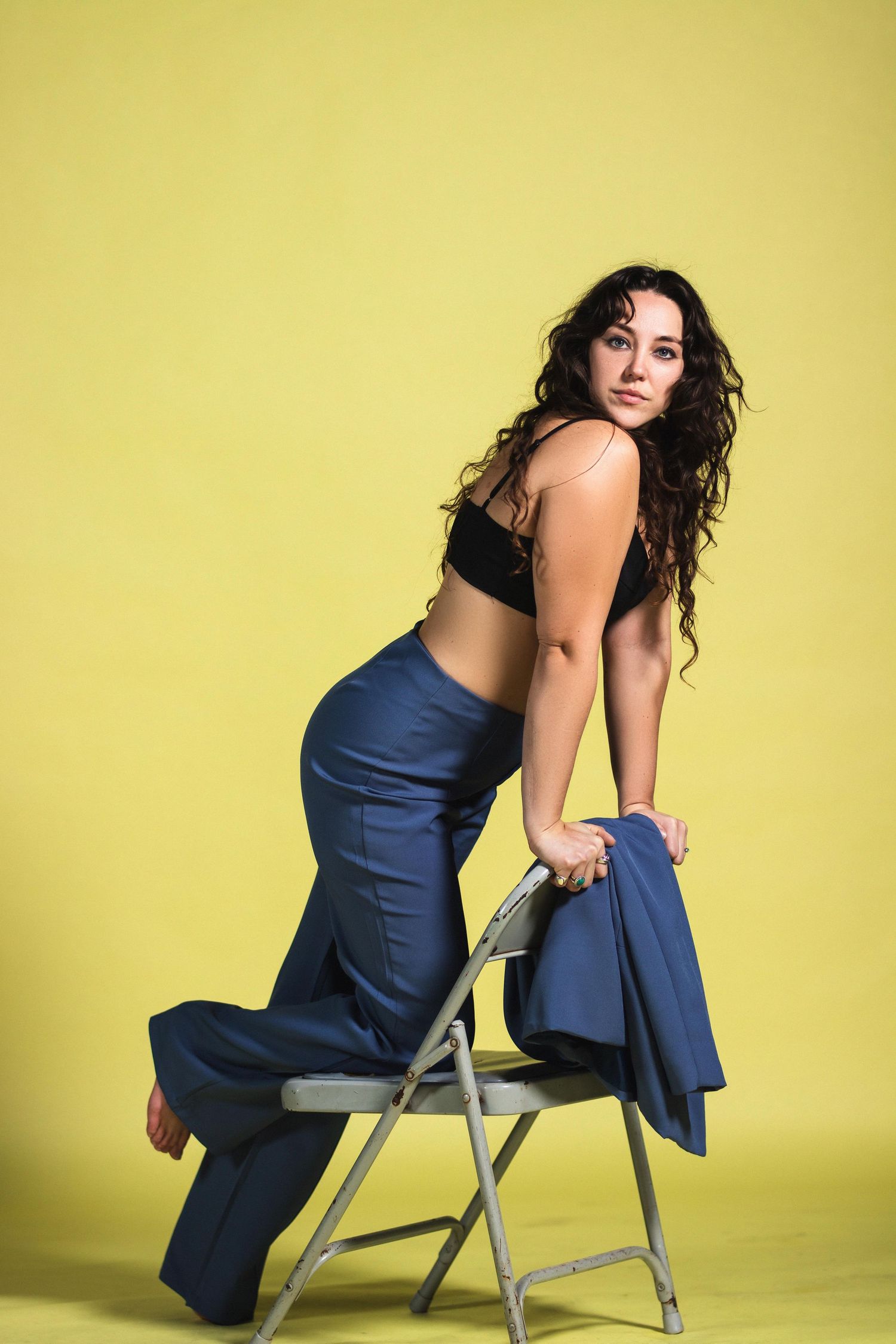 Curly-haired woman in blue wide-leg pants and black crop top posing on folding chair, yellow background.
