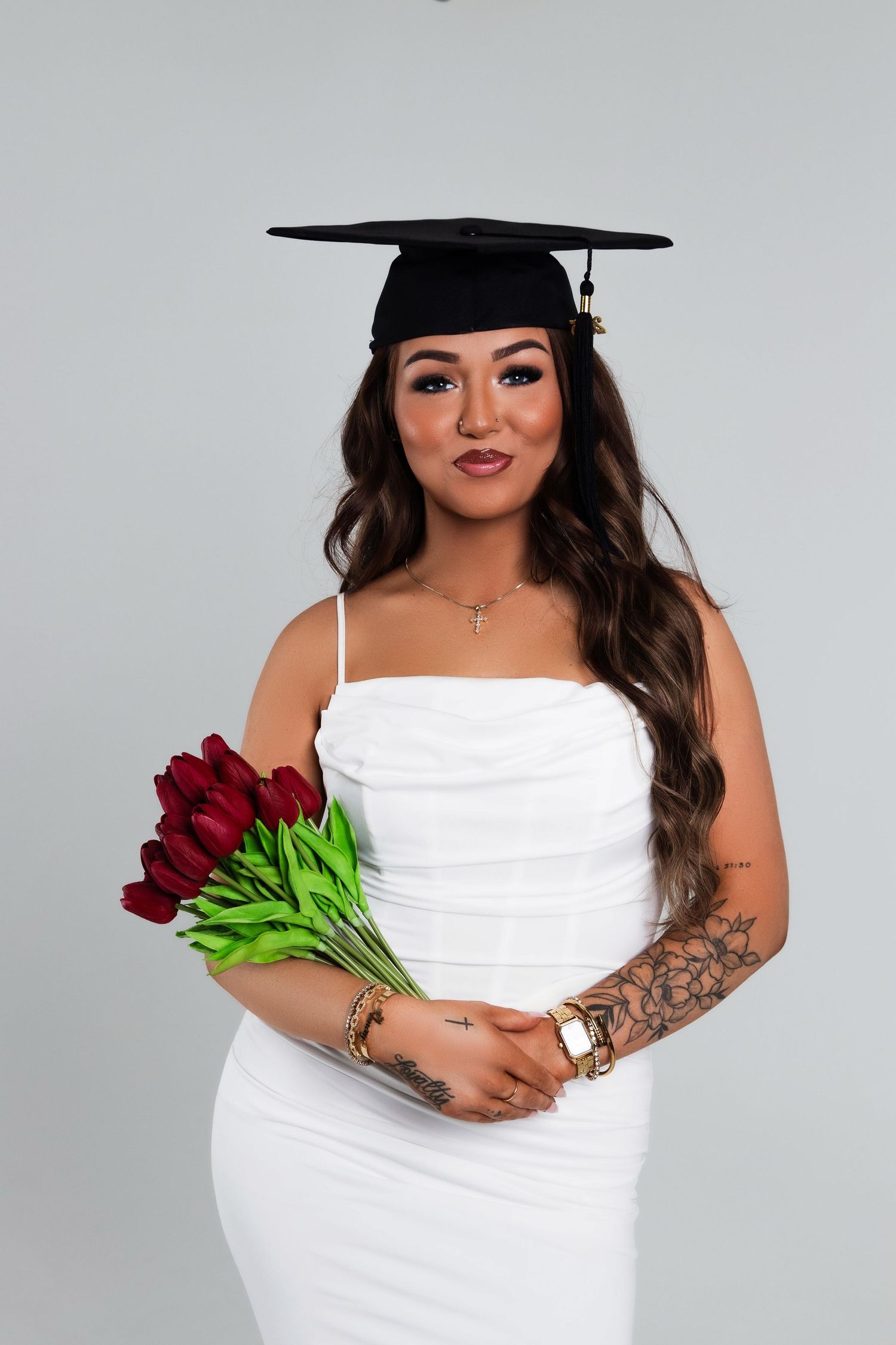 Young woman in white dress and graduation cap holding red tulips with floral tattoo on her arm.