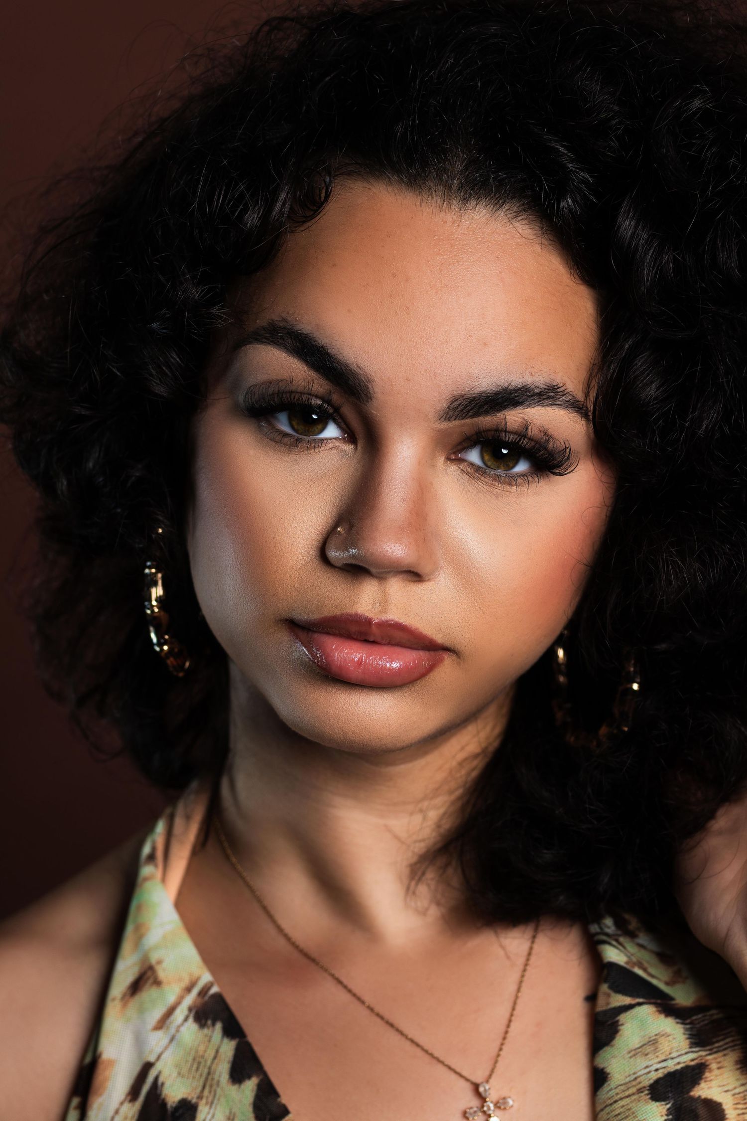 A close-up portrait of a young woman with curly hair, gold earrings, and a necklace on a dark background.