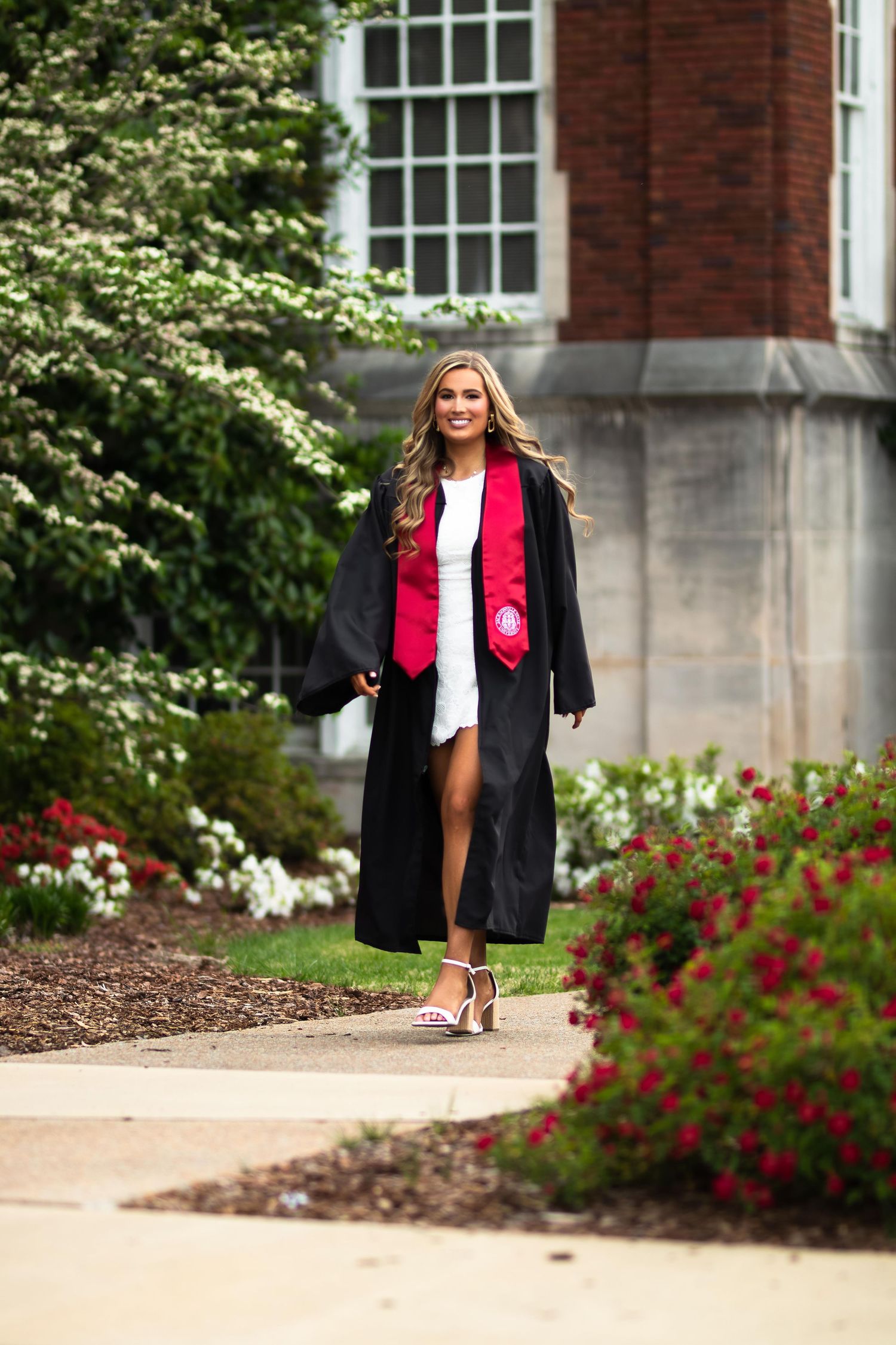 A smiling graduate walks in a black gown and red stole along a garden path beside a brick building.