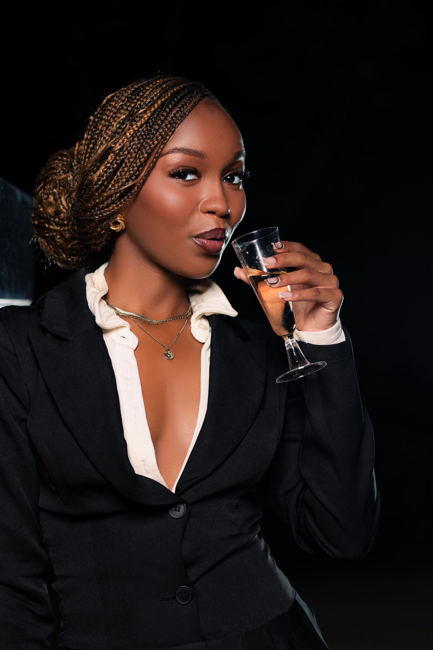 Elegant Black woman in blazer sipping champagne, braided hair, gold necklace, dark background, sophisticated style.