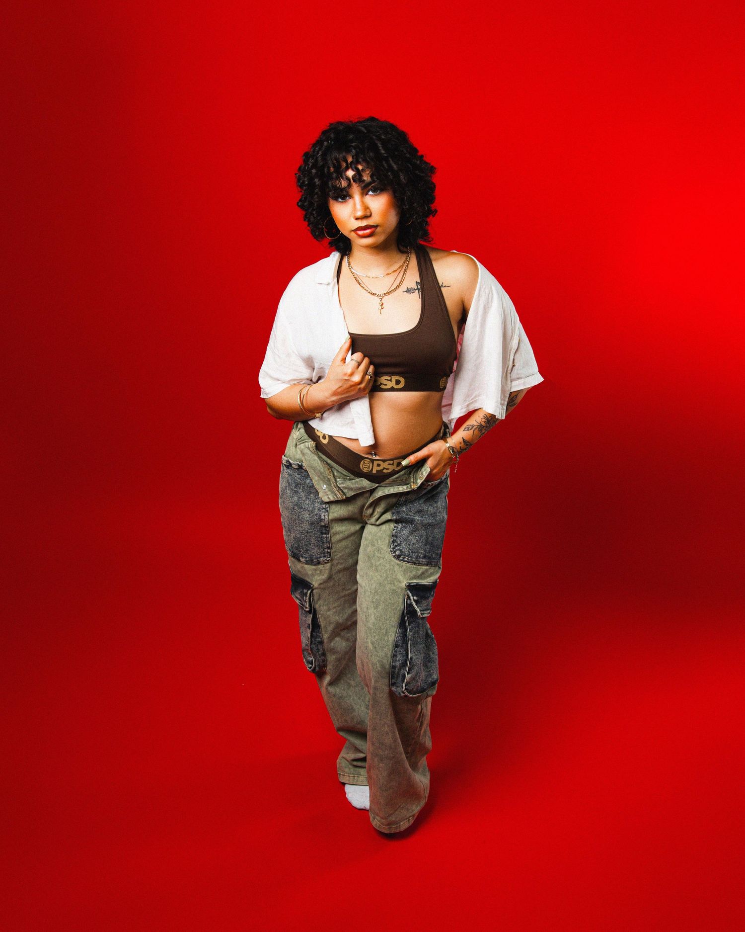 Young woman in brown crop top, white shirt, cargo jeans poses confidently against vibrant red background.