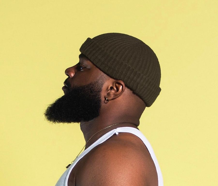 Side profile of bearded man in olive docker beanie and white tank top against yellow background.