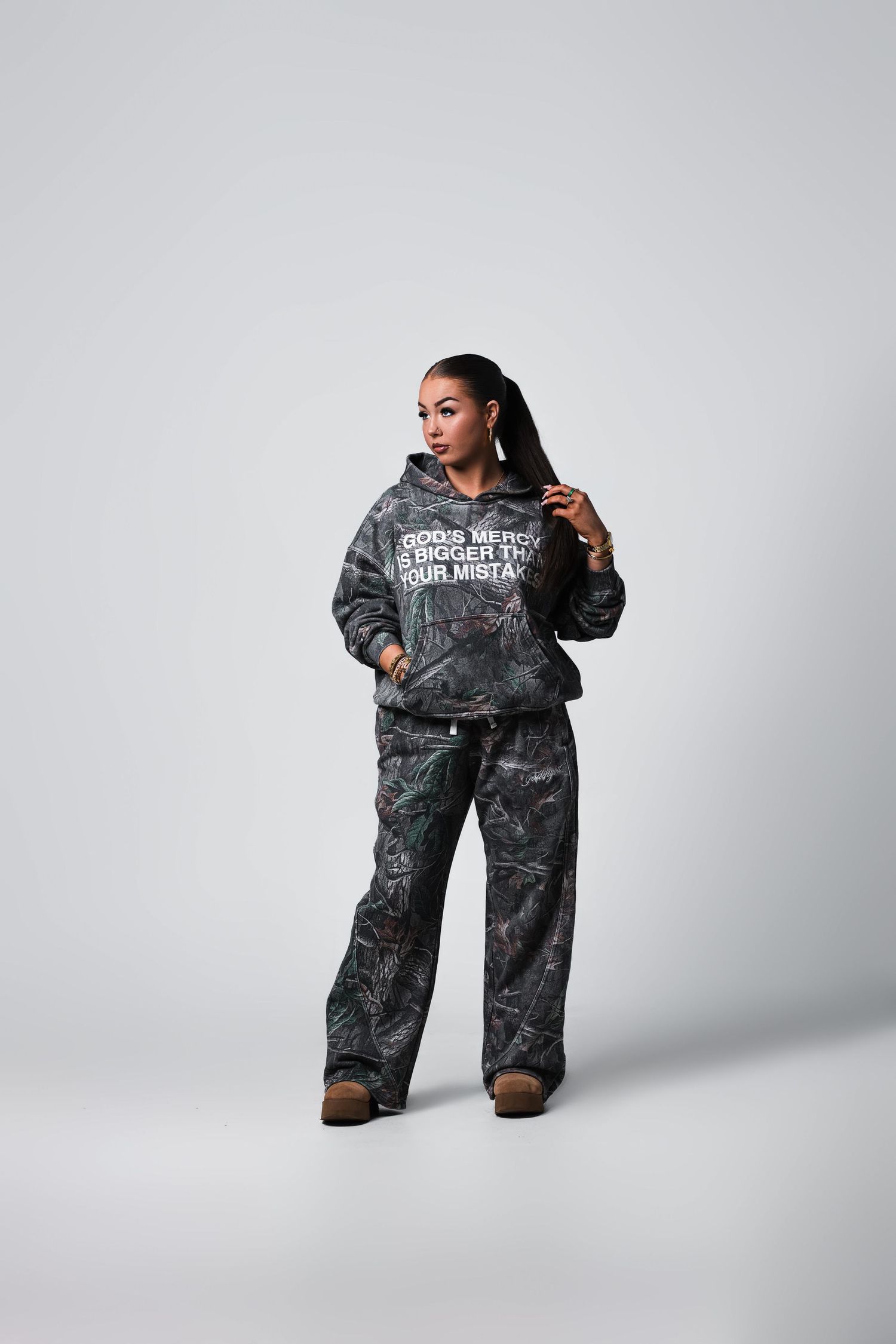 Woman modeling camo tracksuit with graphic hoodie on white background.