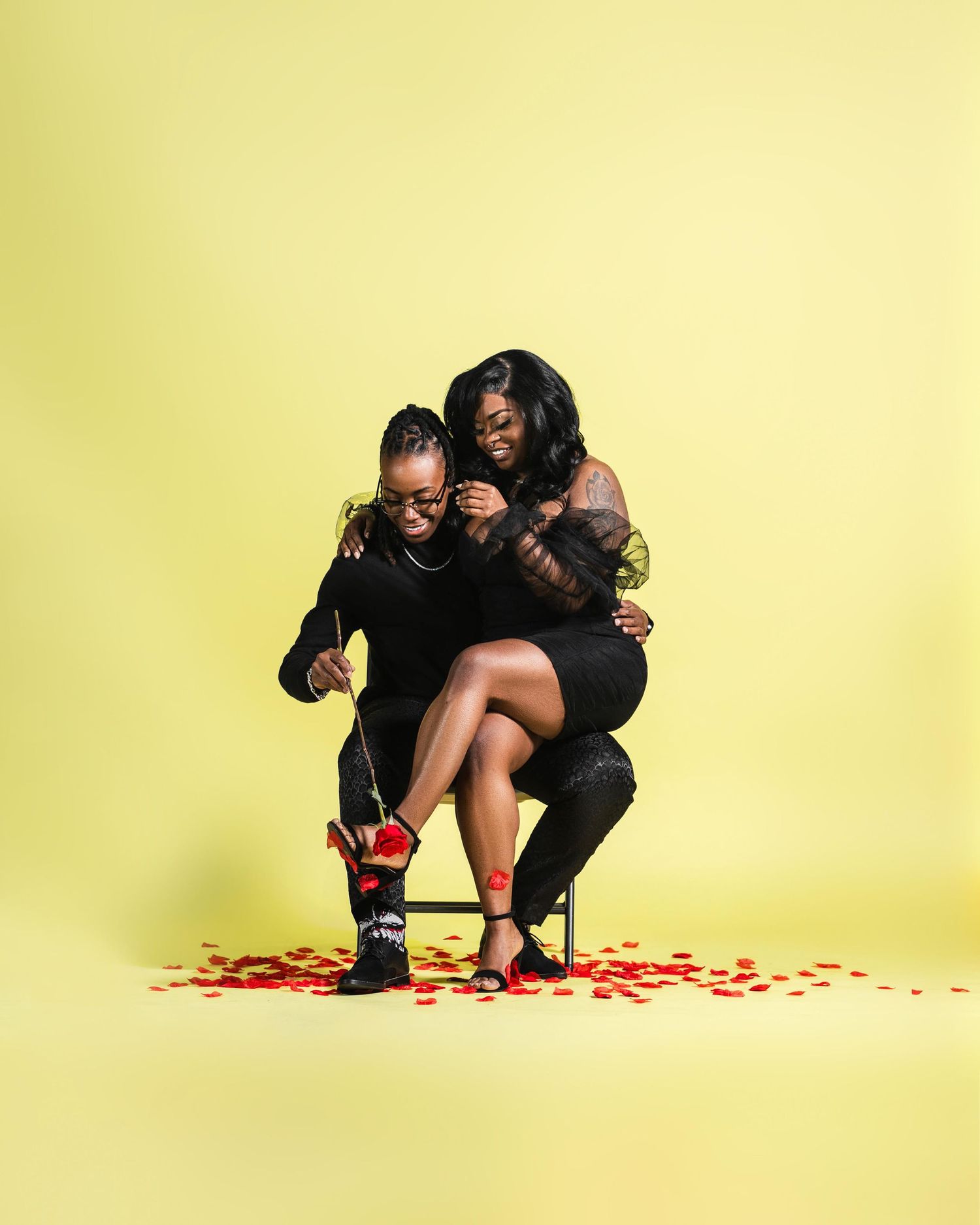 Couple in black outfits laughing together on stool with red rose petals on yellow background.