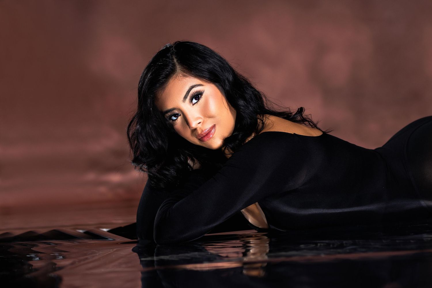 Woman with black wavy hair lies on reflective surface against warm brown studio background.