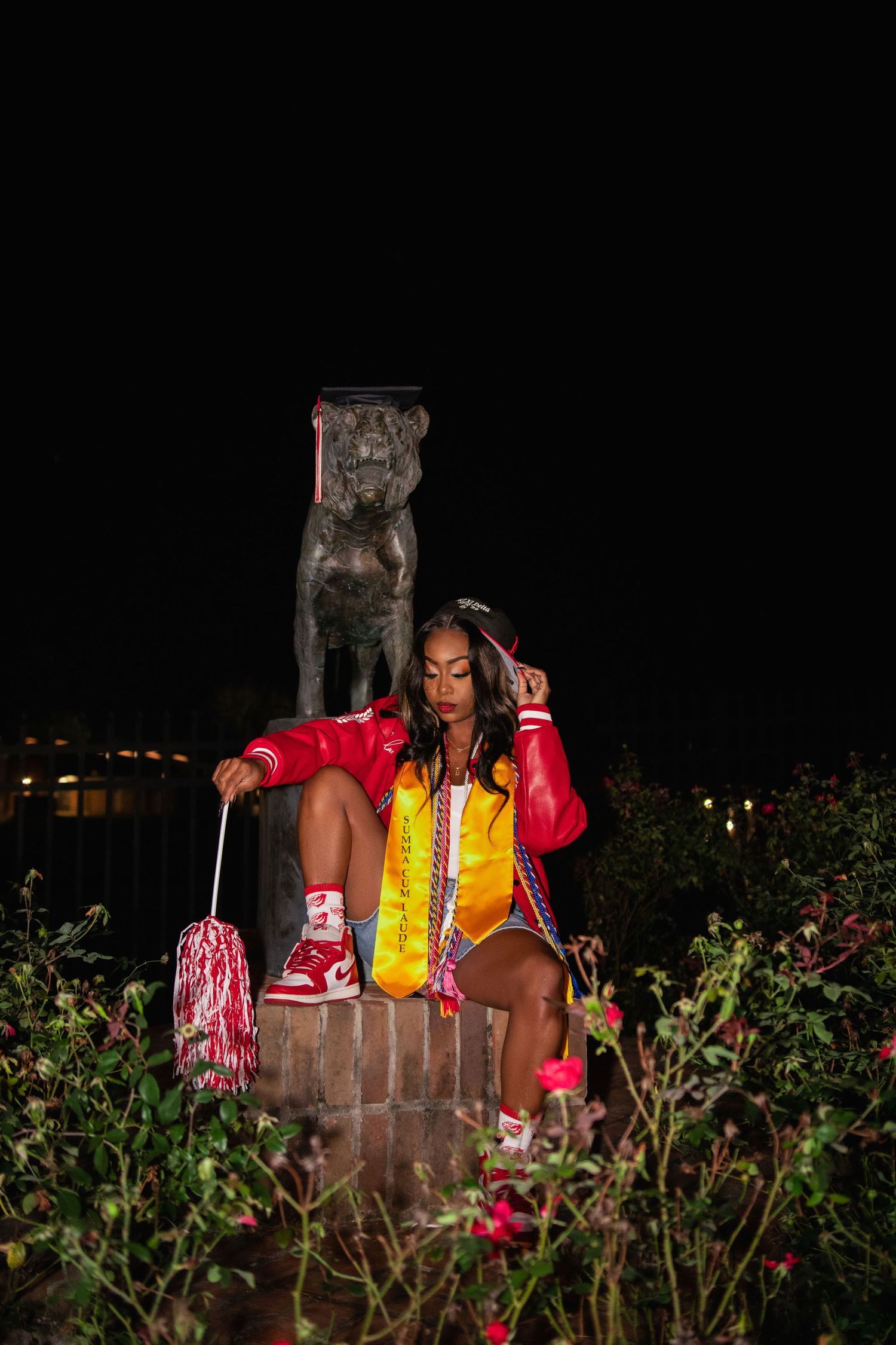 Graduate in red and yellow regalia poses at night beside school mascot statue.
