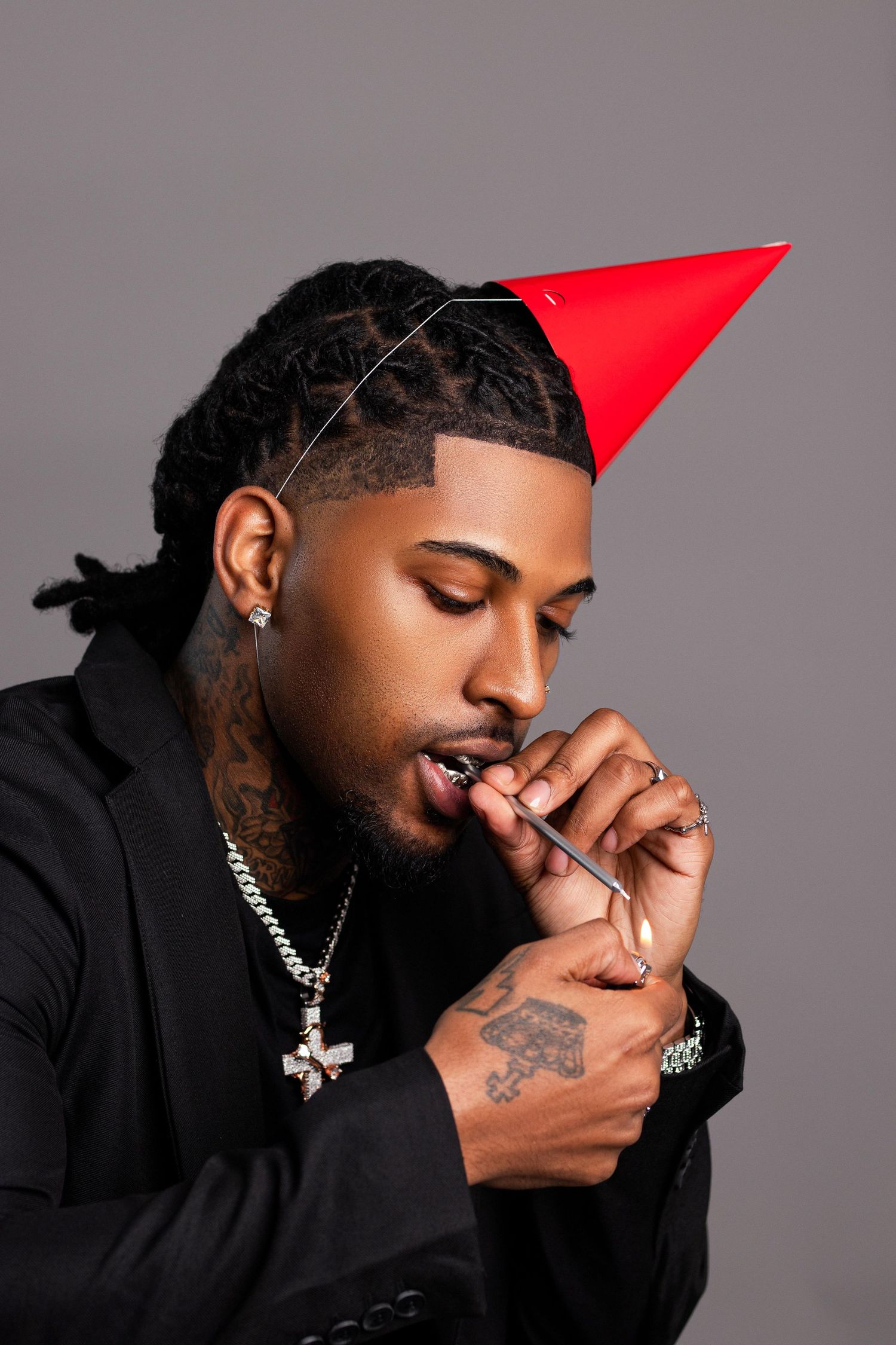 Young Black man with tattoos and silver chain wearing red party hat blowing party horn in black jacket.