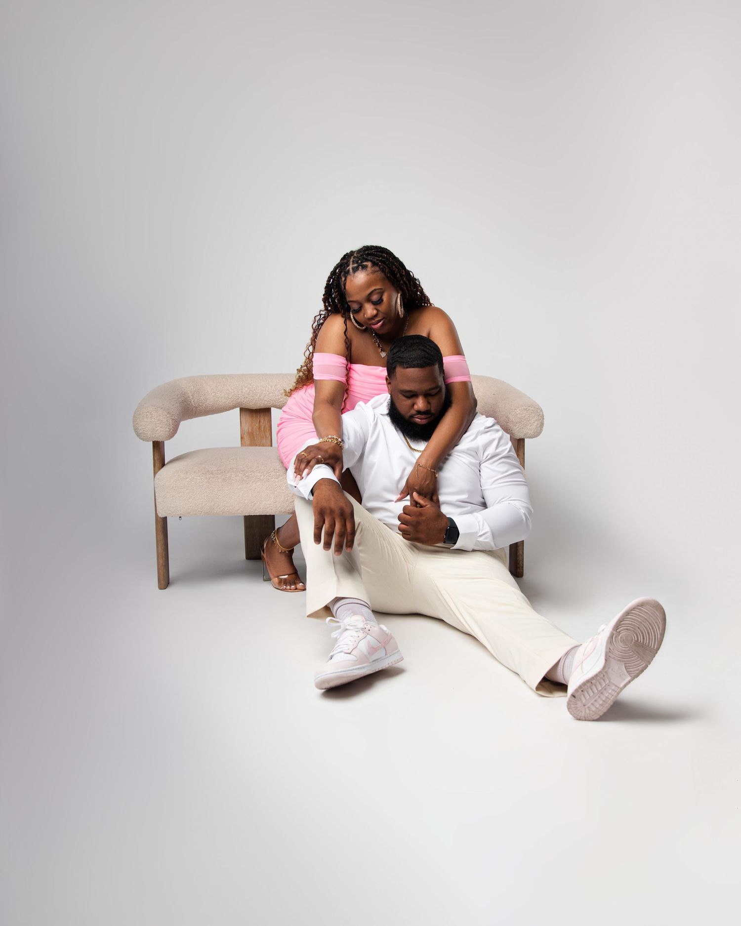 A couple poses elegantly in a studio, woman in pink sits on a beige couch while man in white sits on the floor.