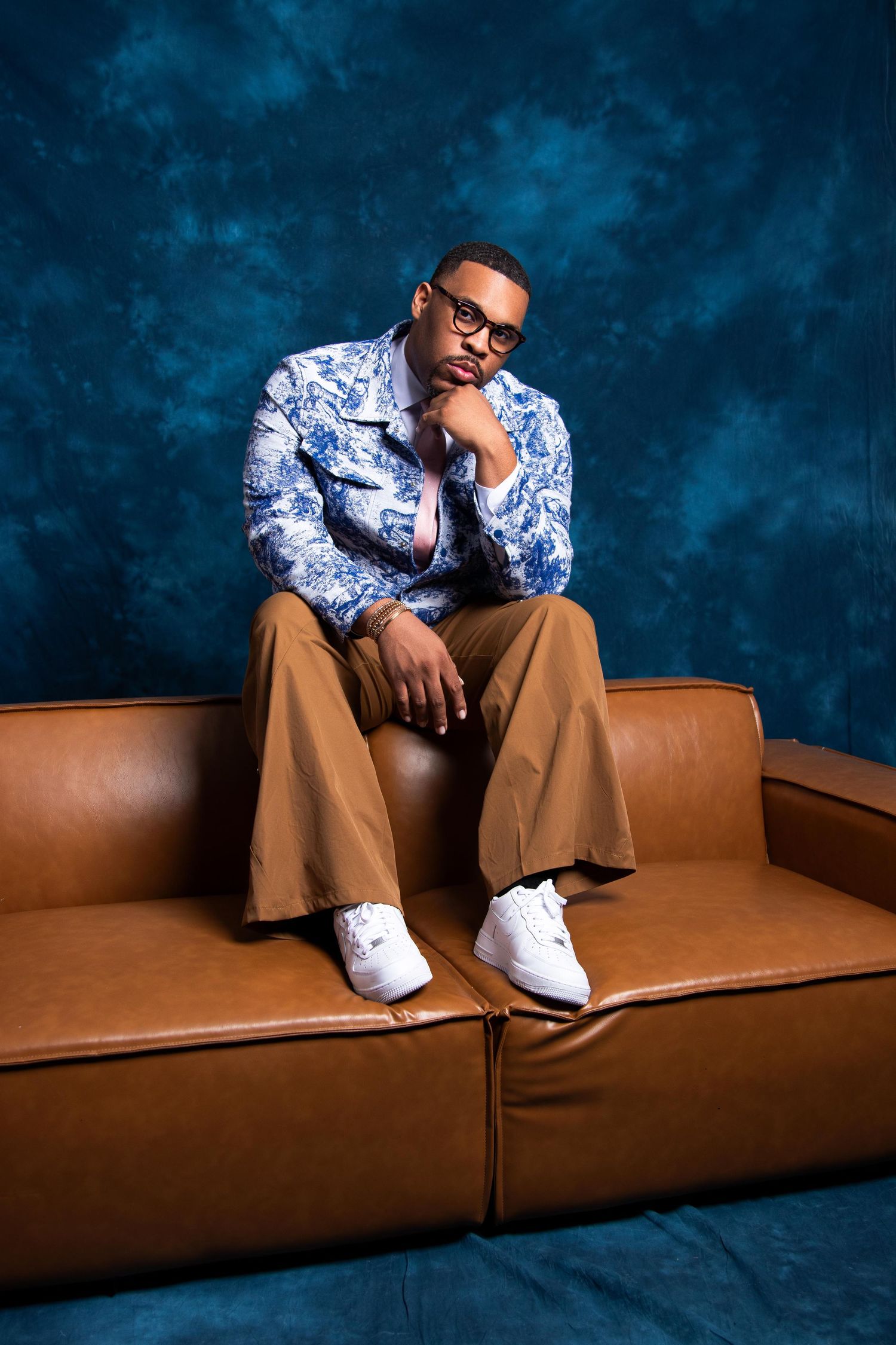 Man in floral shirt, tan pants, and white sneakers sits thoughtfully on a brown leather couch against a blue backdrop.