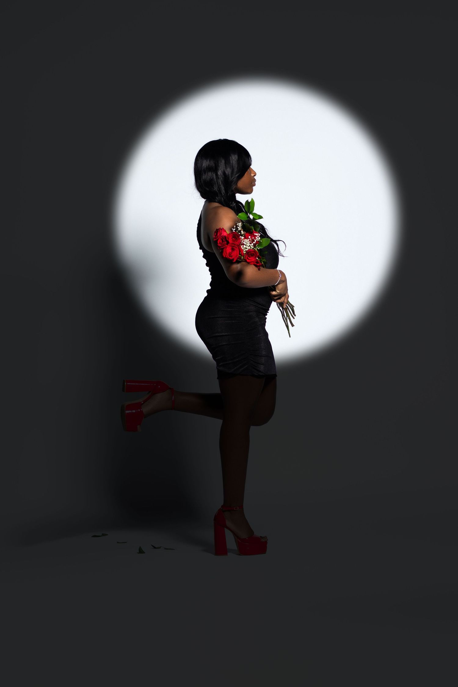 Black woman in black dress holding red roses stands in spotlight against dark gray background.