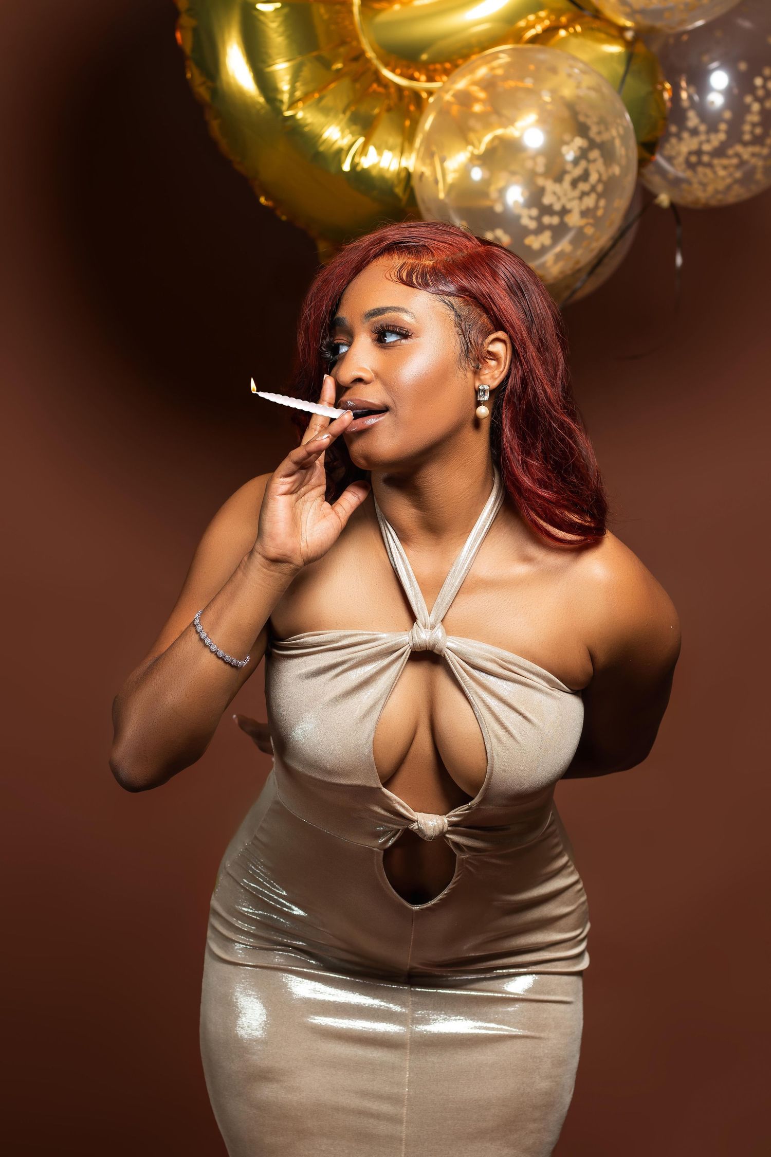 A Black woman with red hair in gold dress smokes a cigarette near gold balloons on brown background.