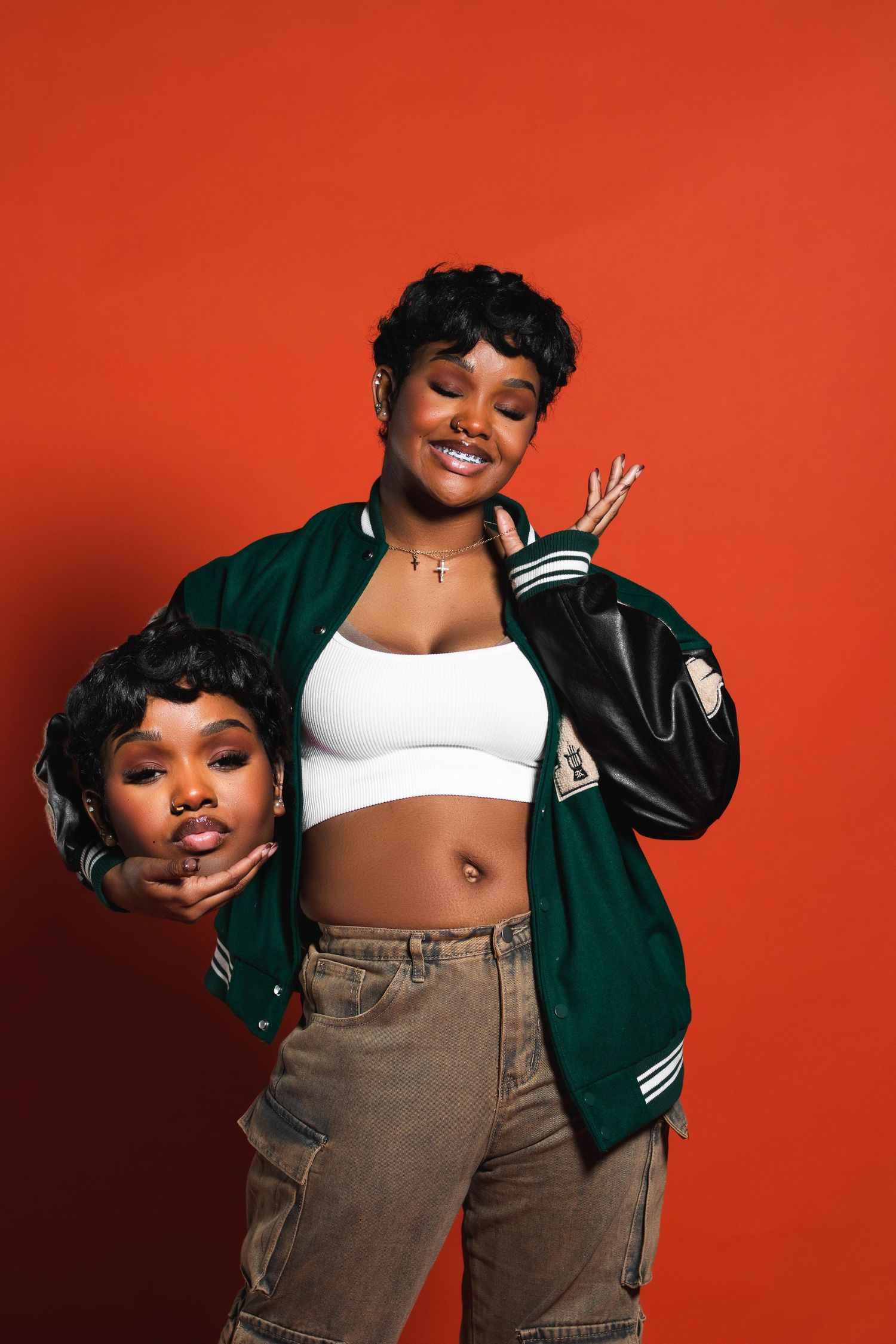 Two Black women pose together, one in green varsity jacket and white crop top on red background.