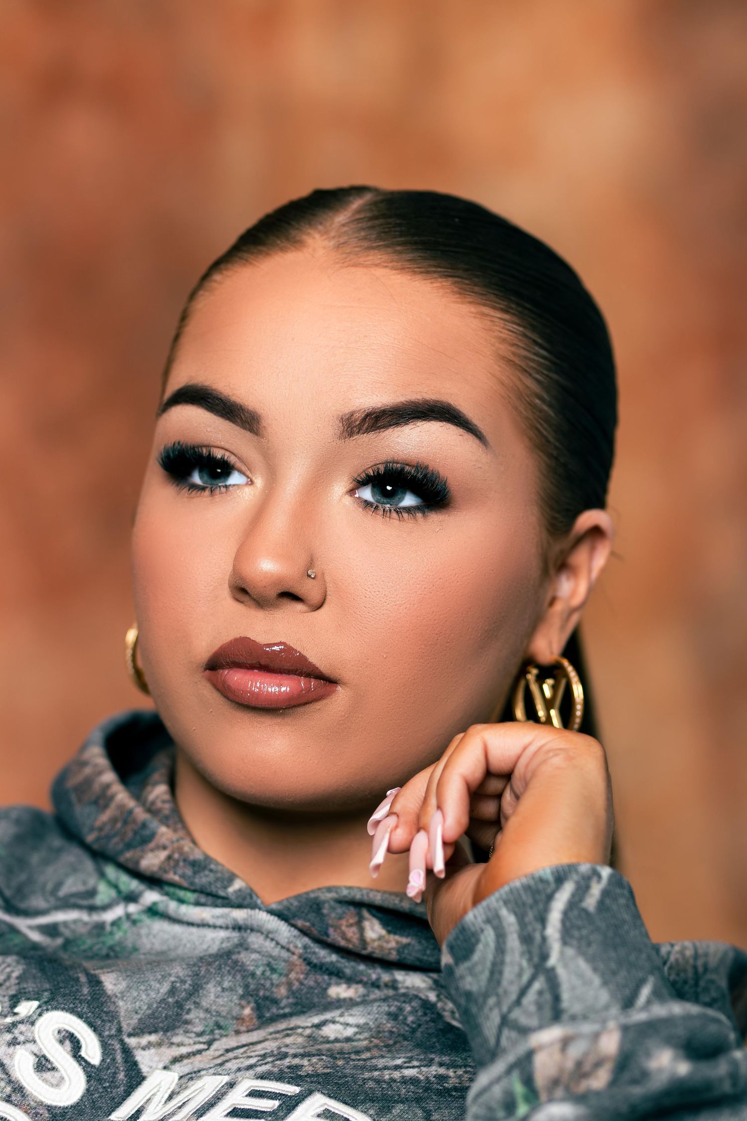 Close-up portrait of woman with blue eyeshadow, nose stud, gold hoop earrings, and camo sweatshirt on brown background.