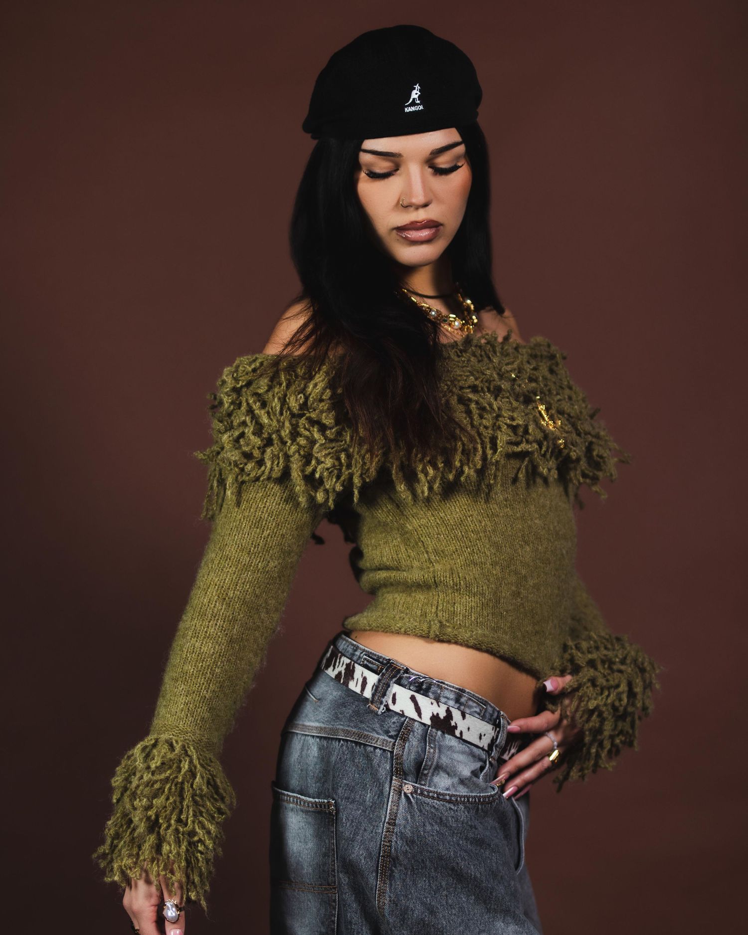 Woman in olive fringe crop sweater and grey jeans wearing Kangol beanie against brown background.