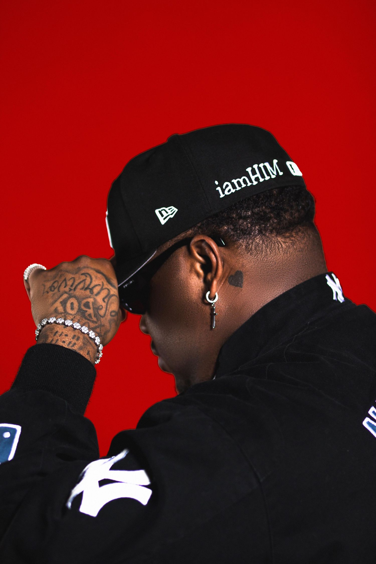 Man wearing a black iamHIM New Era cap and jacket against a vibrant red background.