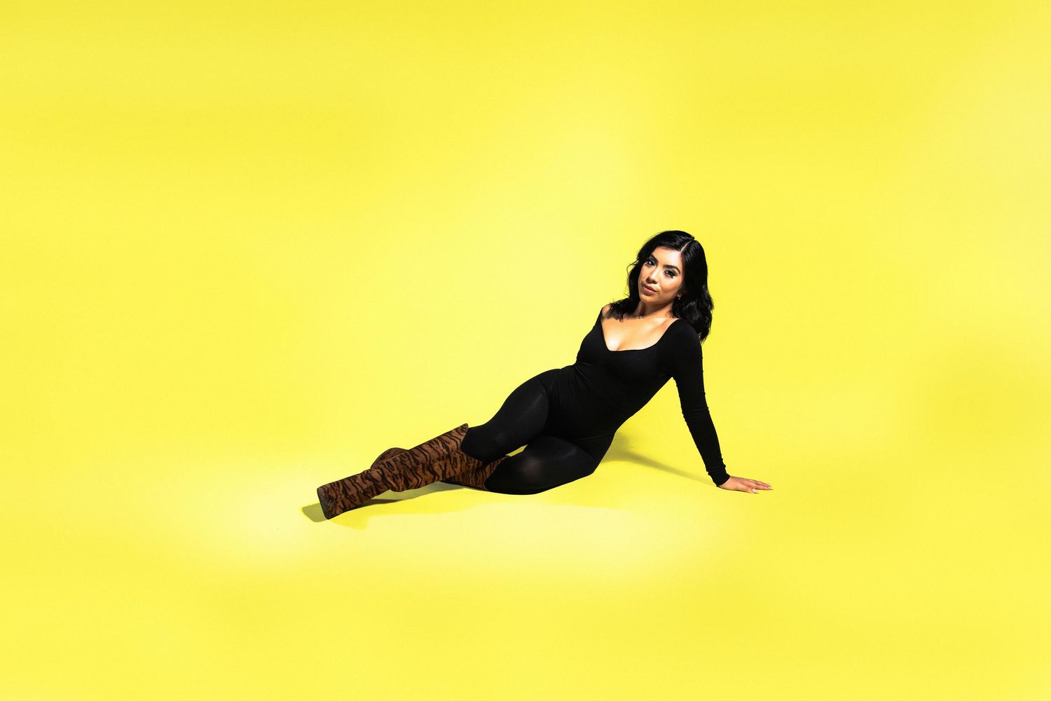 Woman in black bodysuit and brown boots posing on yellow background.